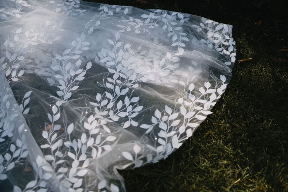 Elegant white floral lace wedding dress fabric with delicate leaf embroidery spread on green grass.