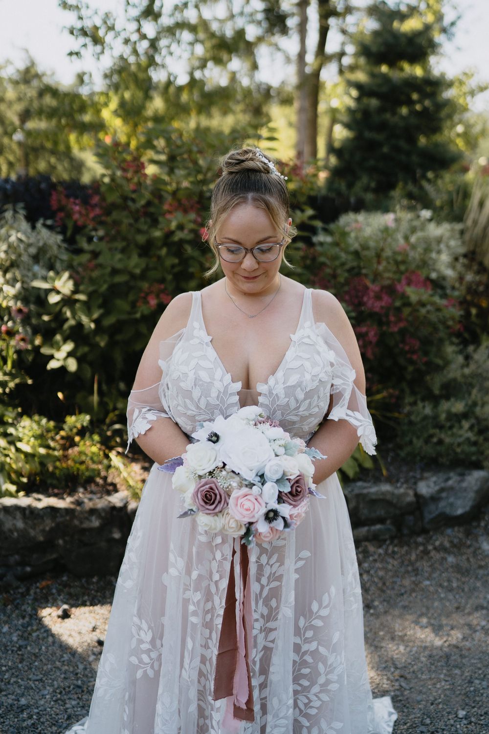Bride in lace wedding gown holding white and pink floral bouquet with ribbon, standing in garden.
