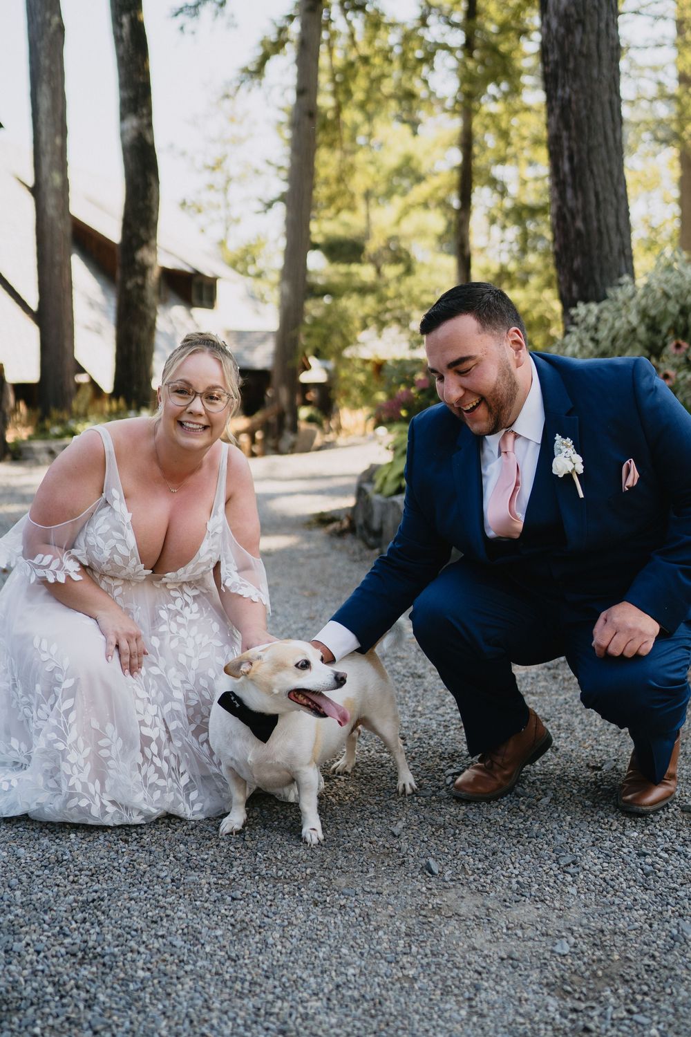Bride and groom crouching on gravel path petting their small dog wearing a bow tie at outdoor wedding venue.