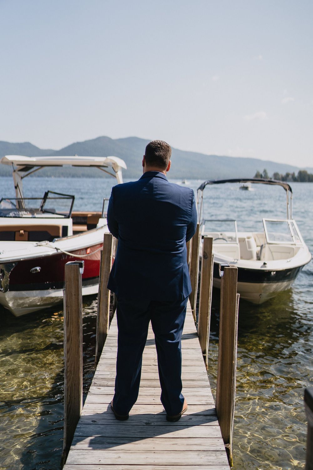 Man in navy suit standing on wooden dock overlooking lake with boats and mountain backdrop.