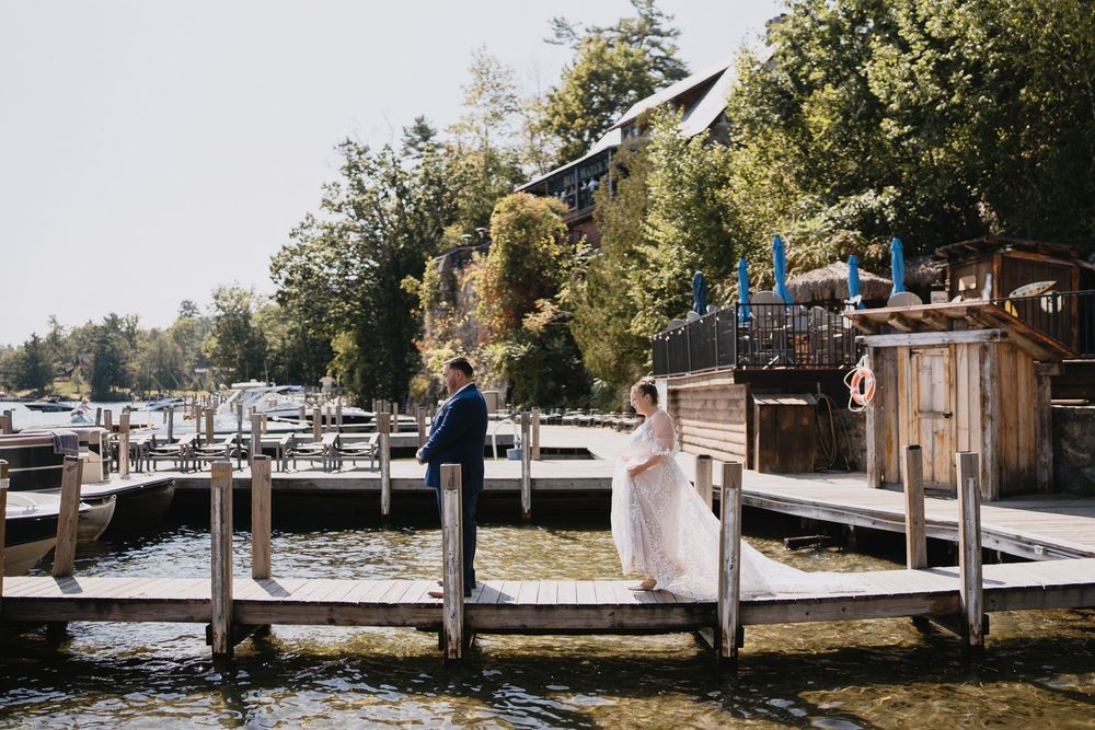 Bride and groom stand on a wooden dock by a lake, surrounded by lush trees and a waterfront resort on a sunny day.