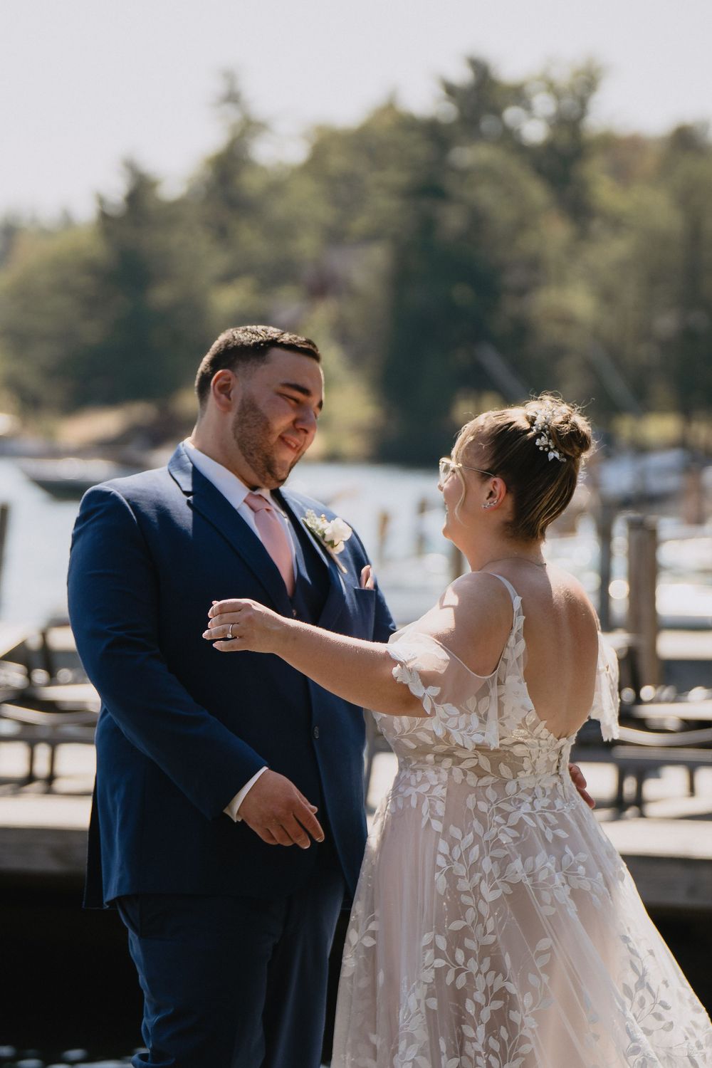 A bride and groom share a tender moment on a lakeside dock, she in lace gown, he in navy suit with pink tie.