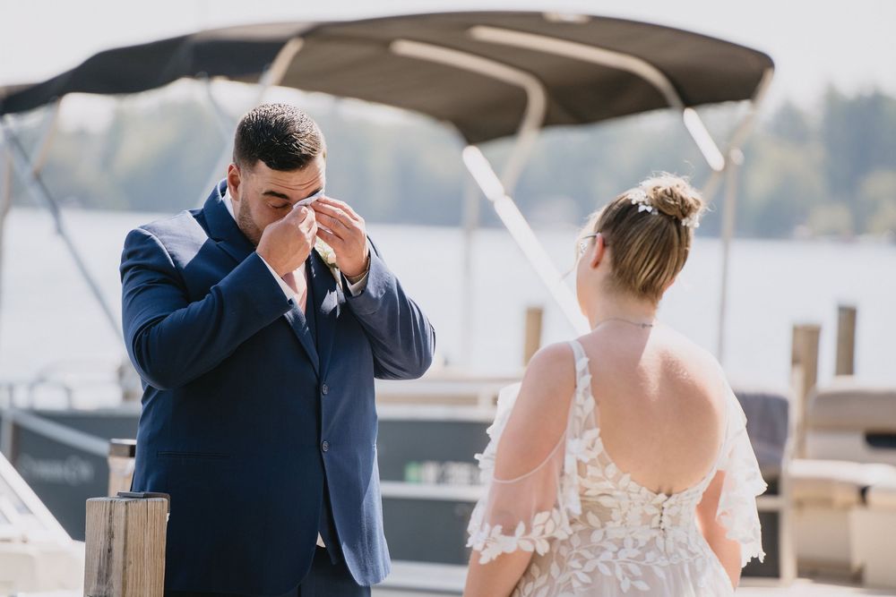 Emotional groom wiping tears during outdoor waterfront wedding ceremony with bride in lace dress.