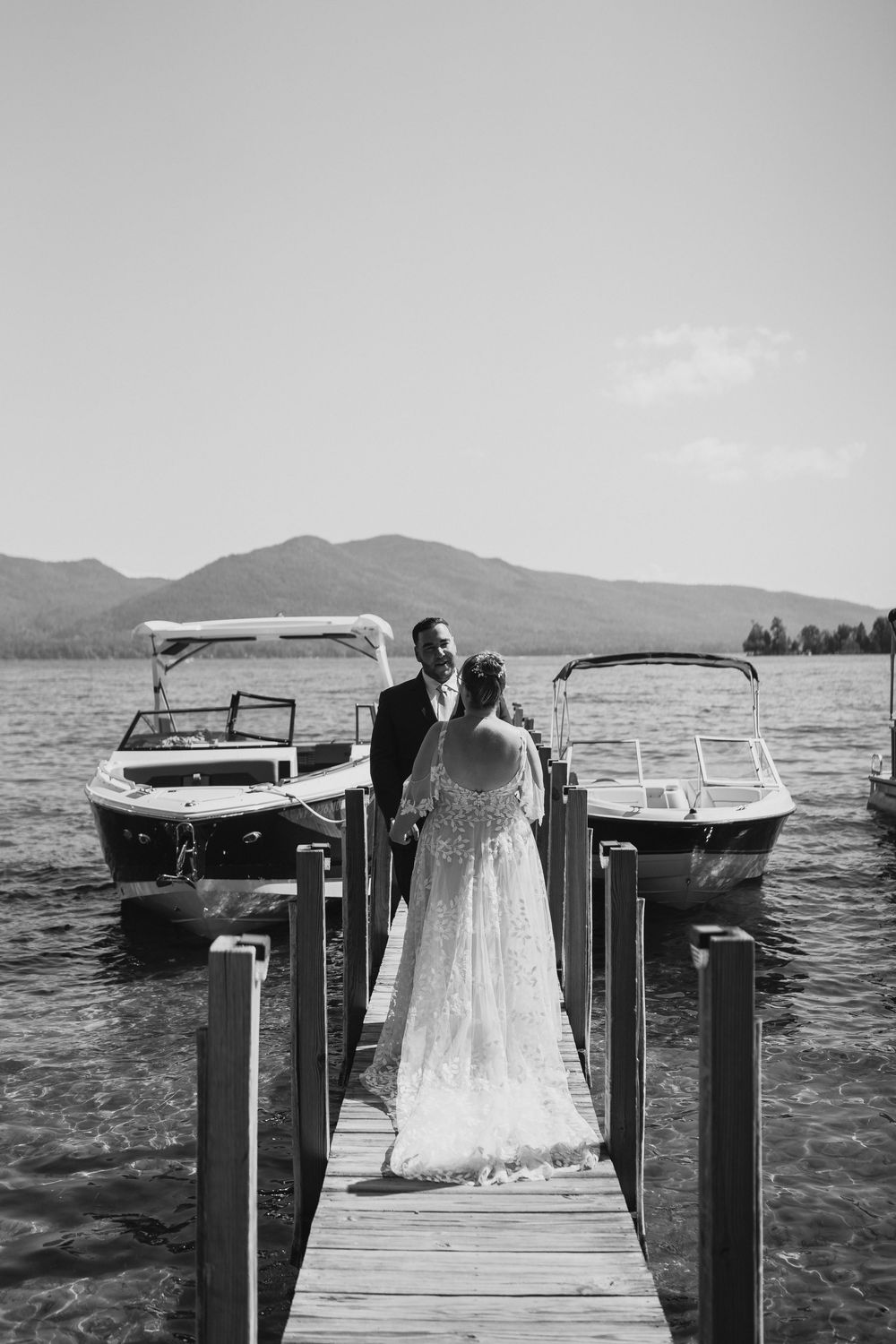 Black and white photo of a bride and groom standing on a lake dock with boats and mountains in the background.