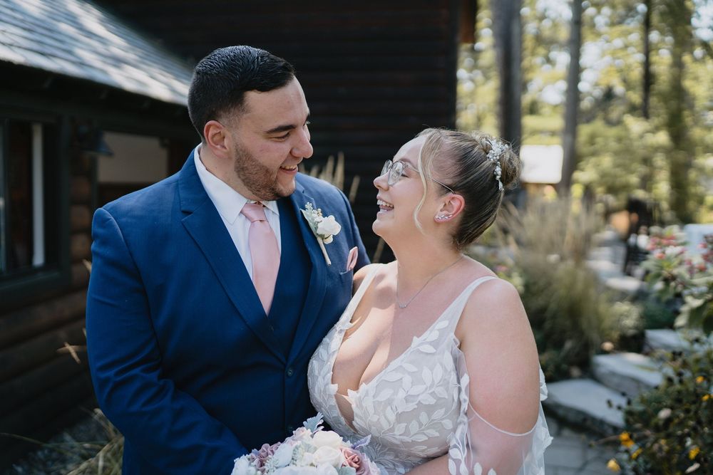 A bride and groom share a romantic moment outdoors, smiling at each other, groom in blue suit with pink tie.