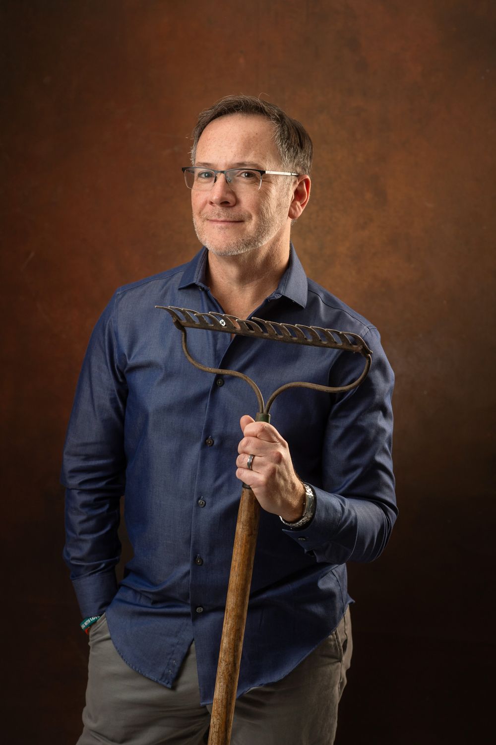 Middle-aged man with glasses holding a vintage garden rake, wearing a blue shirt, posed against a brown studio background.