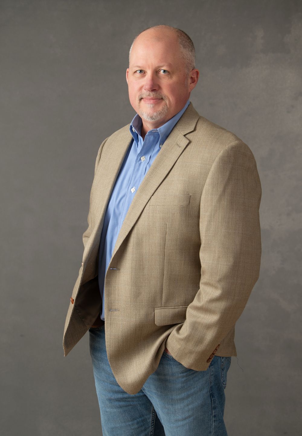 Middle-aged bald man in beige blazer and blue shirt posing confidently with hands in pockets against gray background.