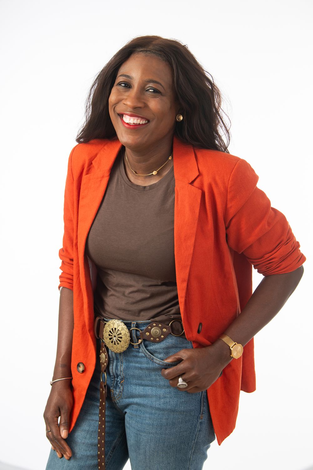 Smiling Black woman in orange blazer, brown top, jeans, and decorative belt buckle posing confidently on white background.