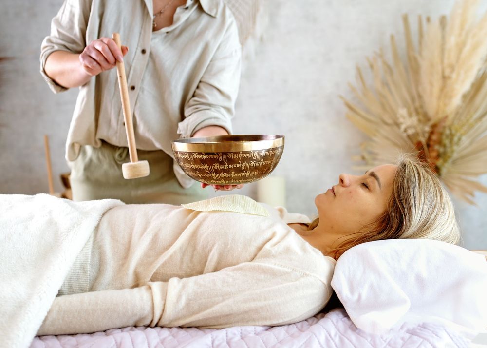 Therapist performing a Tibetan singing bowl sound healing therapy session on a relaxed woman lying on a massage table.