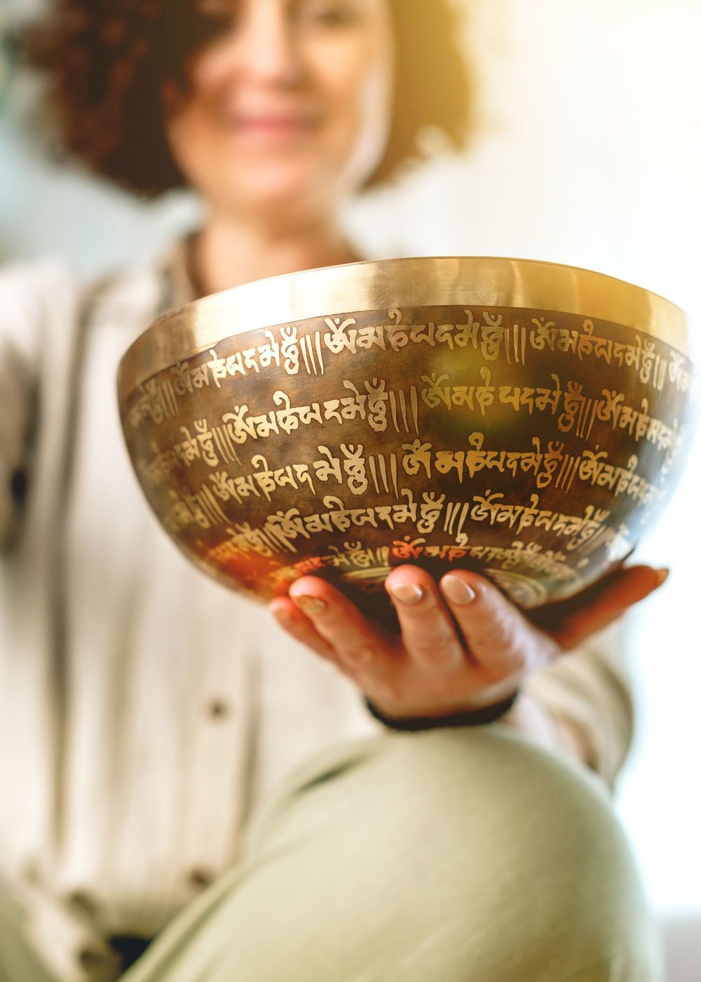 Woman smiling while holding a gold Tibetan singing bowl engraved with Sanskrit mantras during a meditation session.