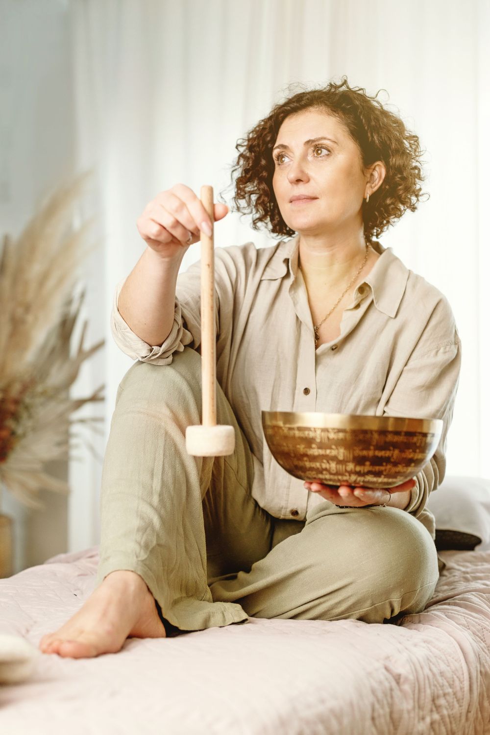 Woman sitting cross-legged on a bed holding a Tibetan singing bowl and mallet in a serene, sunlit room.