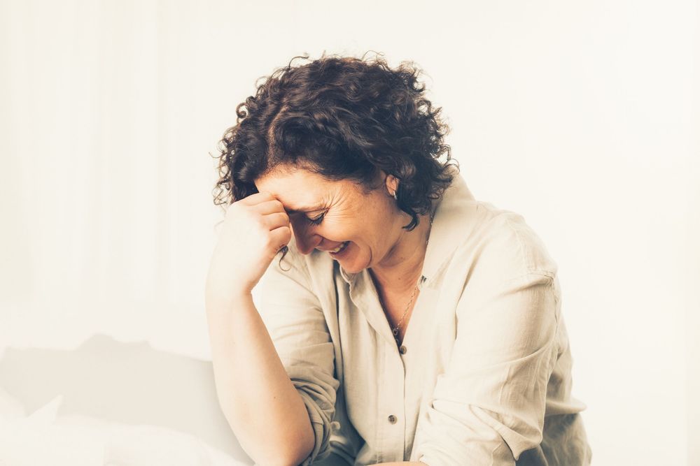 A stressed woman with curly hair holds her forehead in her hand while sitting down, looking overwhelmed.