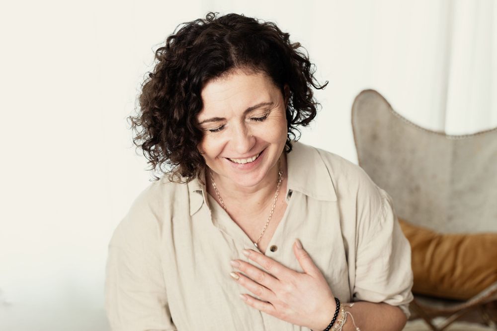 A person with curly dark hair wearing a beige shirt smiles warmly while touching their chest in a bright room.