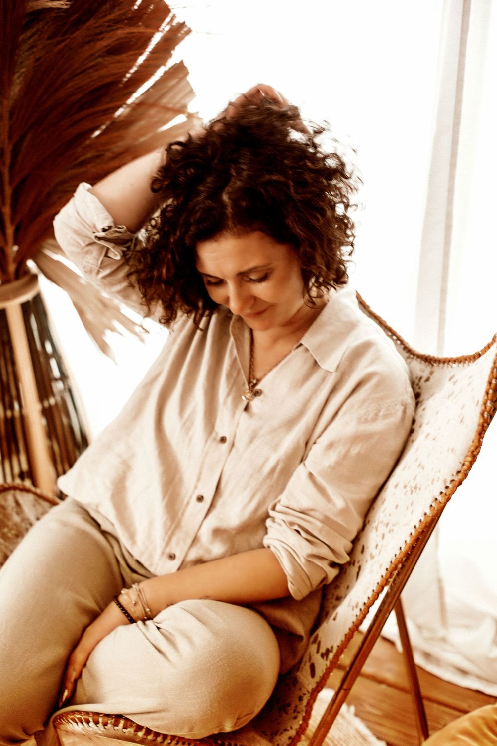 A woman with curly hair sits relaxed in a wicker chair, wearing a linen shirt, smiling softly in warm natural light.
