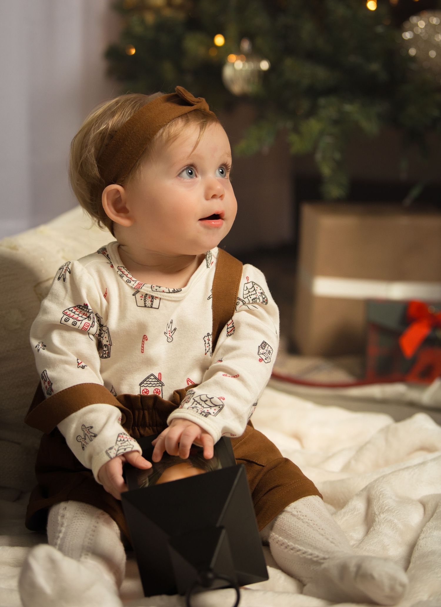 Adorable baby in Christmas pajamas sitting near a decorated holiday tree holding a gift box.
