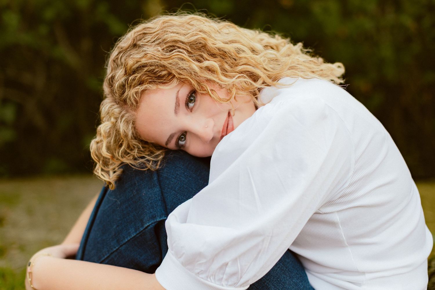 A Kansas City senior sits with her knees up, arms wrapped around her legs and head resting on her knees, smiling softly at the camera.