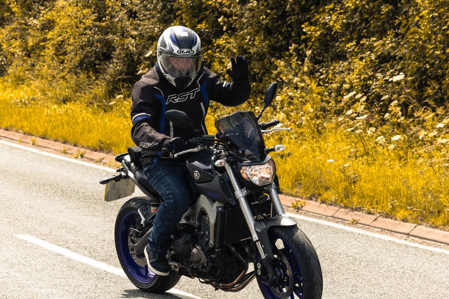 Motorcyclist in helmet and gear riding a black motorcycle on a road surrounded by lush green and yellow foliage.