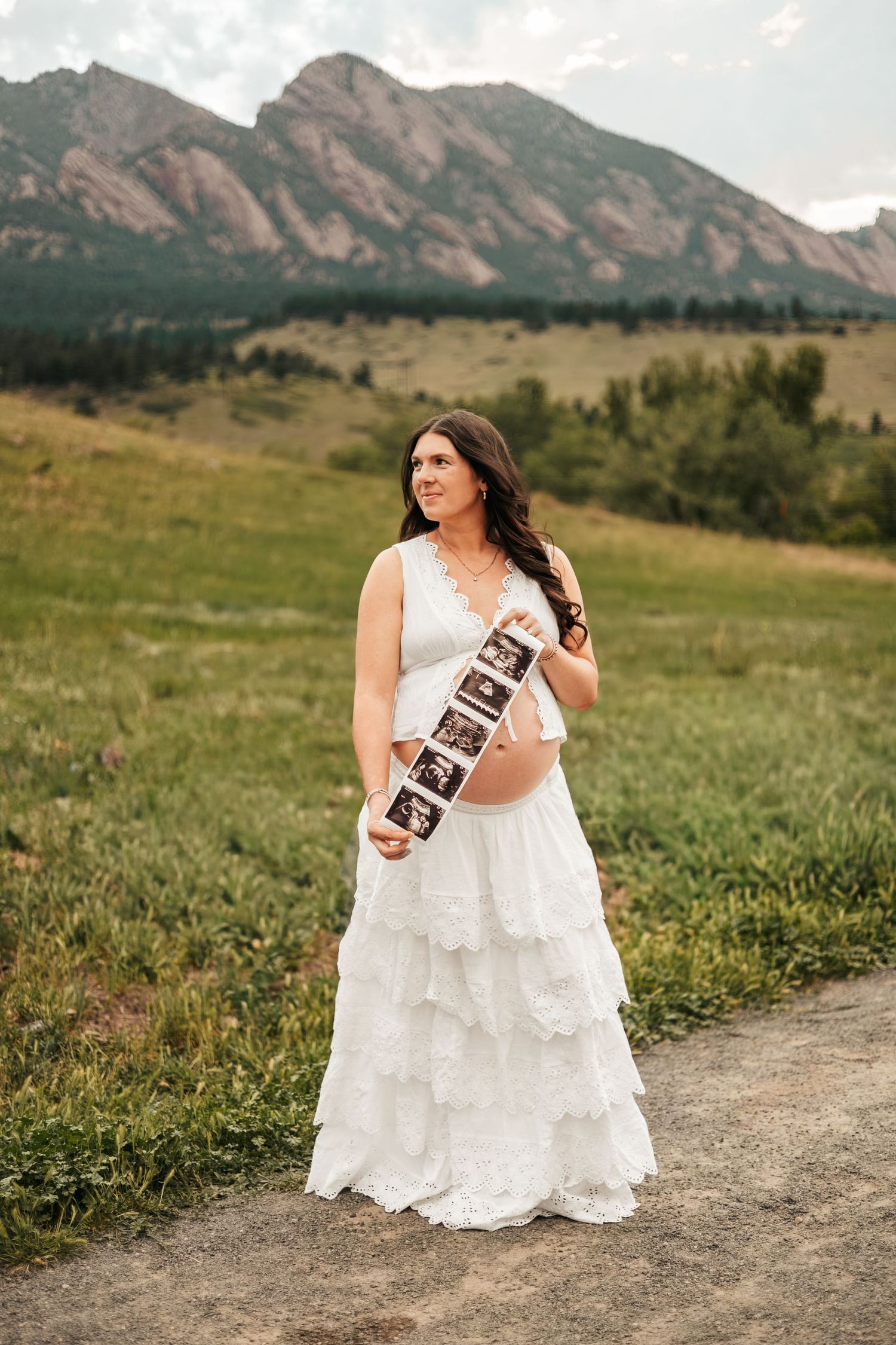 Pregnant woman in white lace dress holds ultrasound photos at Colorado Flatirons maternity photoshoot| Boulder Family Photographer