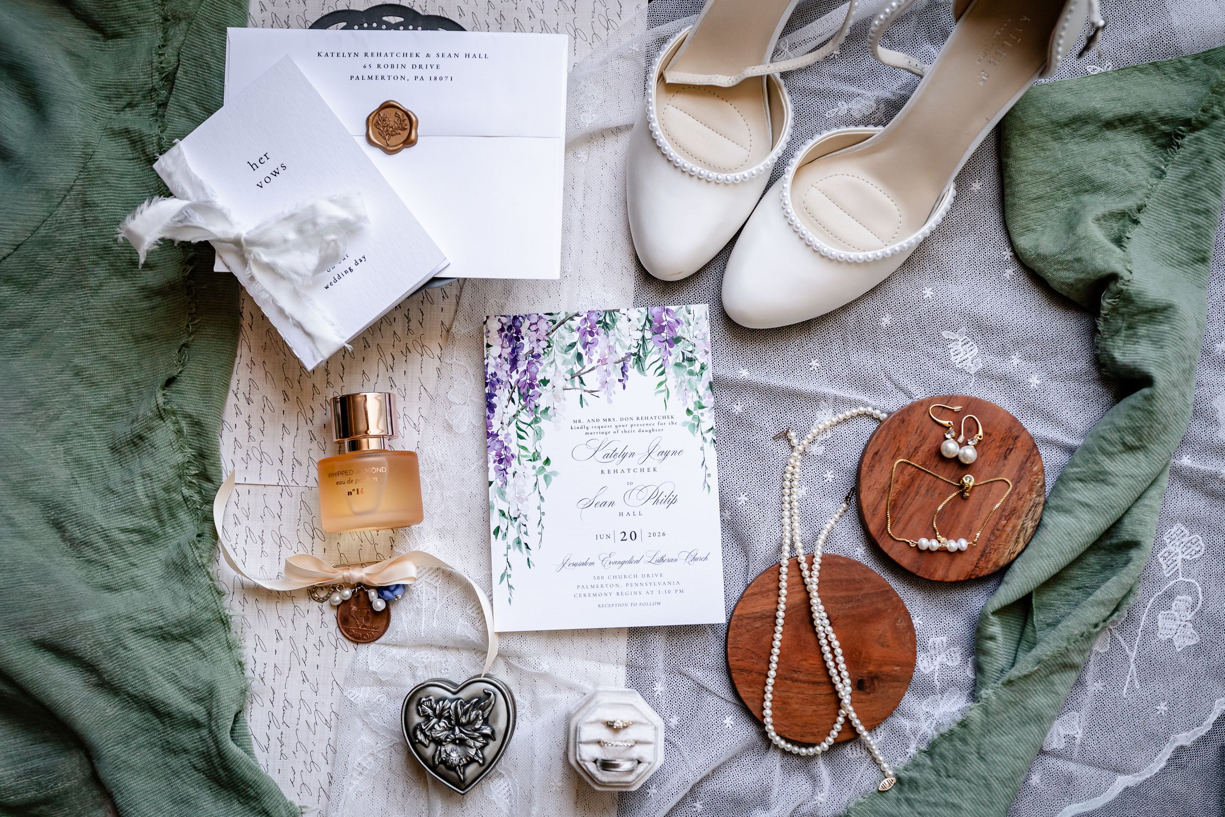Flat lay of wedding invitation, bridal shoes, perfume, jewelry, and accessories on green fabric.