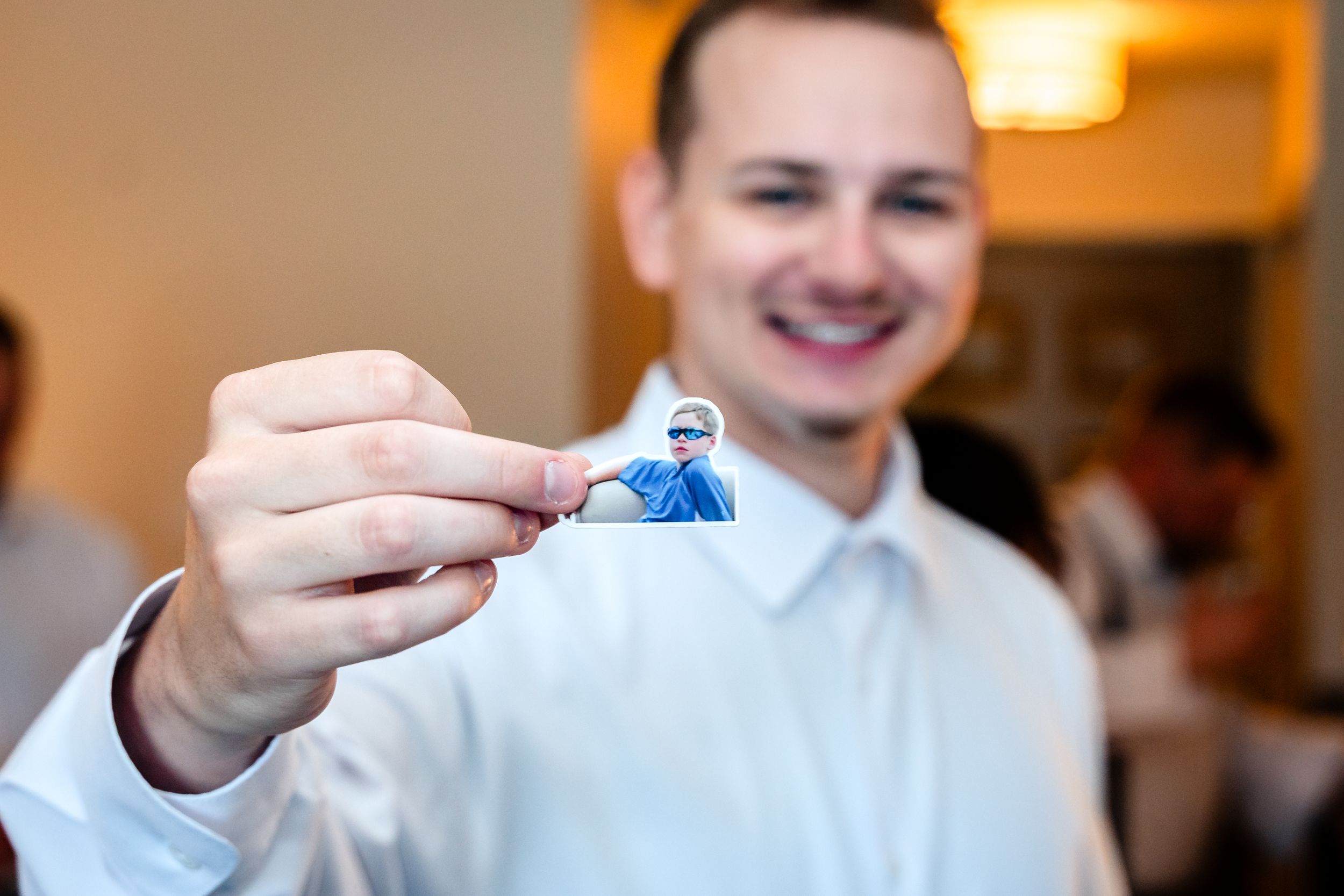Smiling groom in white shirt holding small blue wedding figurine at reception venue.