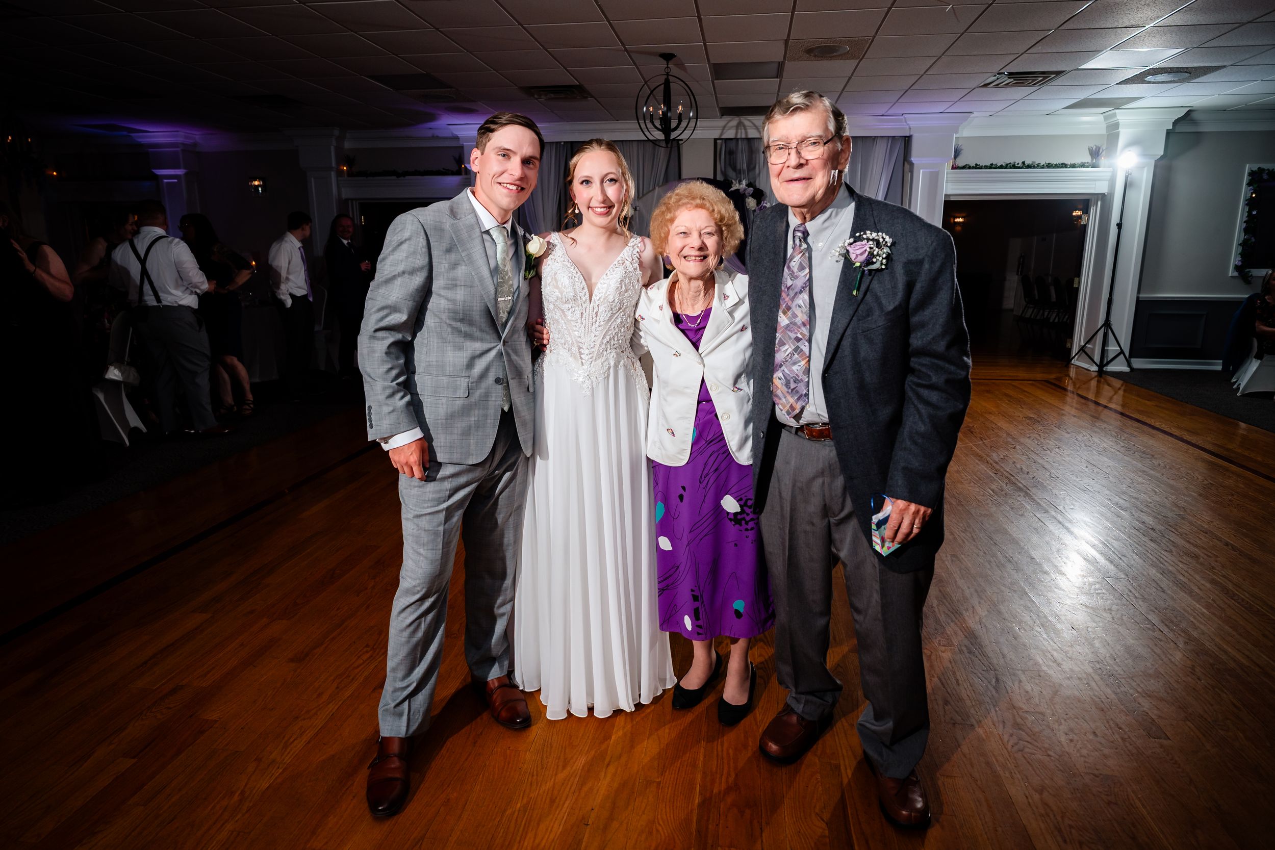 Four people posing together at a wedding reception on a dance floor, including a bride in white and an older couple.