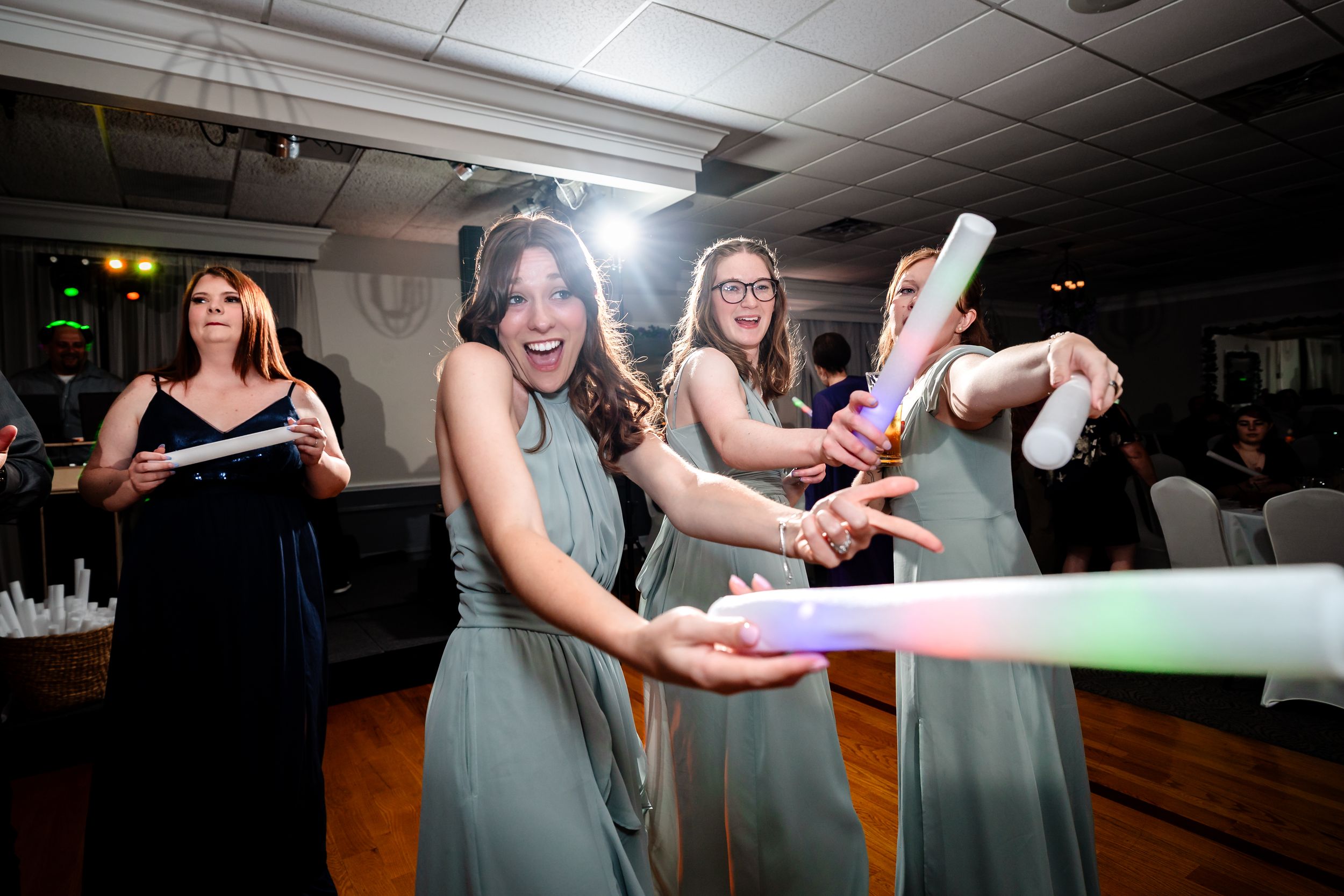 Bridesmaids in sage green dresses celebrate with colorful glow sticks at wedding reception.