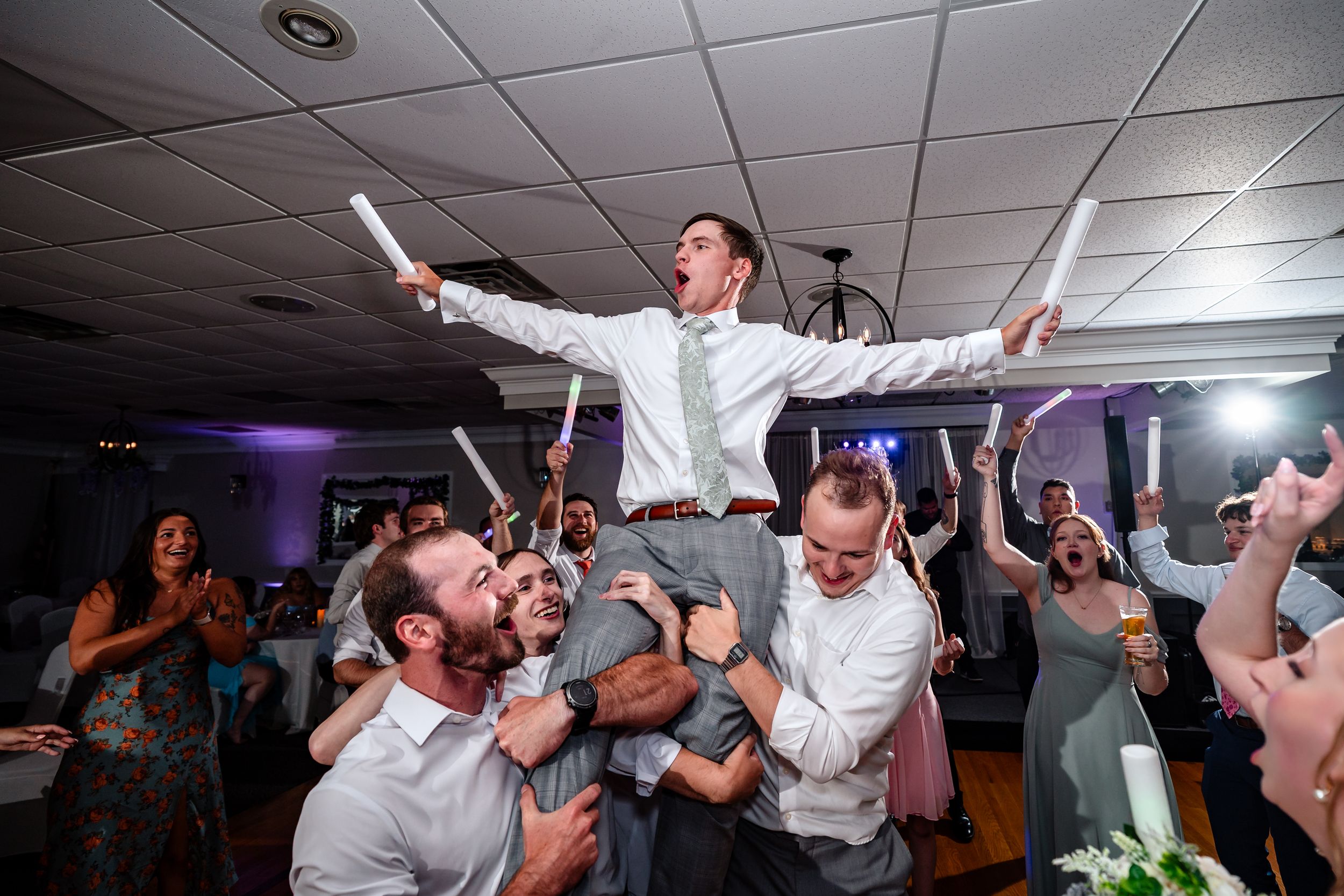 Groom lifted by groomsmen on shoulders during lively wedding reception dance floor celebration with glow sticks.
