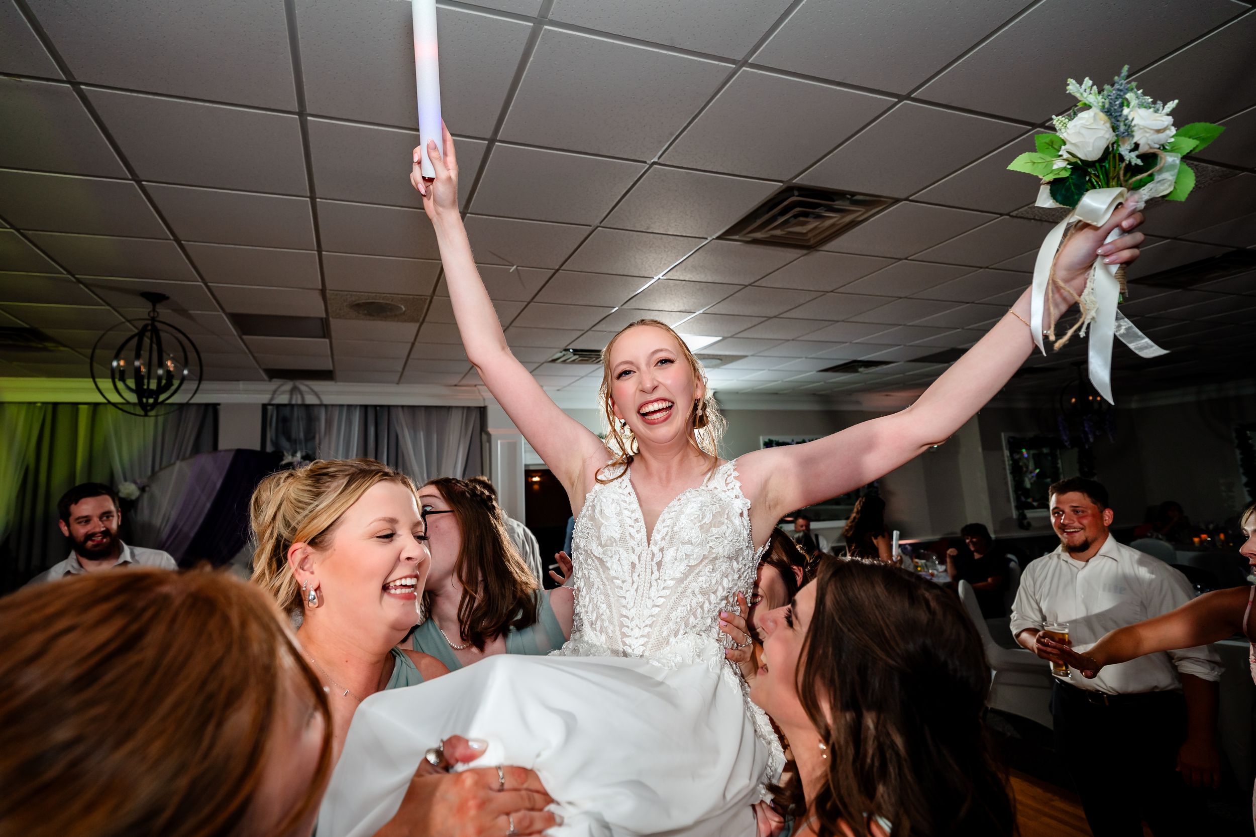 Bride lifted by bridesmaids on shoulders holding bouquet aloft during joyful wedding reception celebration.