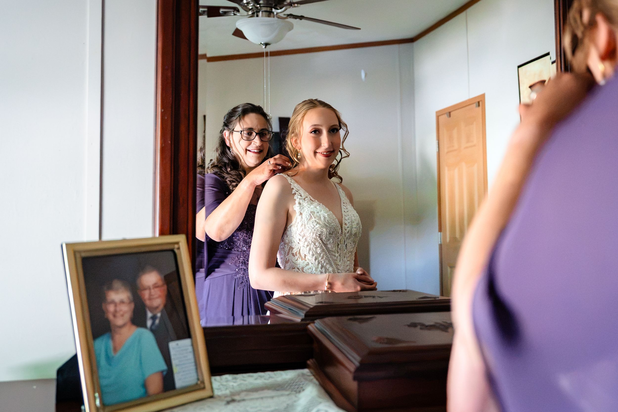 A bridesmaid helps a bride with her wedding dress in front of a mirror during bridal preparations.