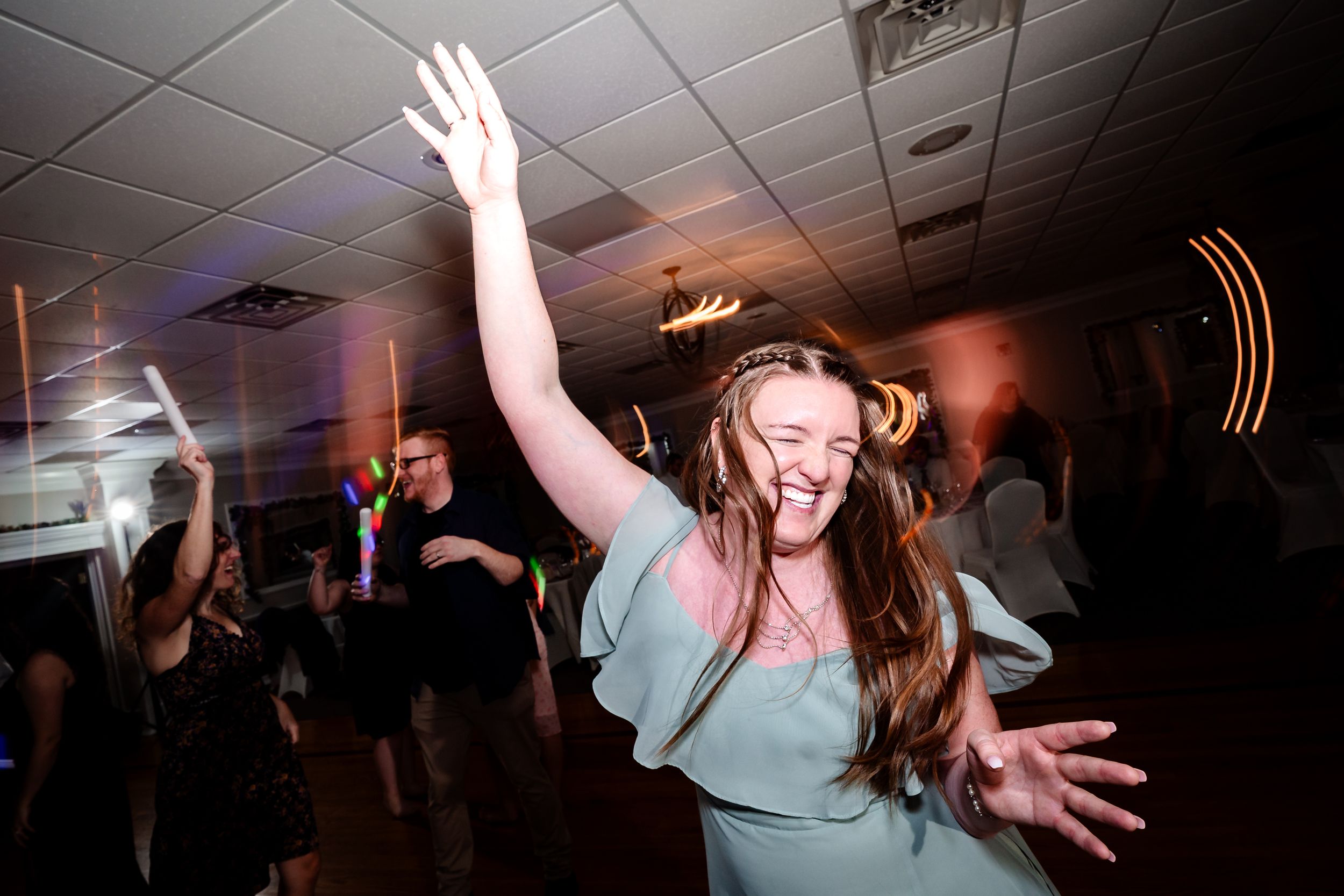 A joyful woman in a sage green dress dances with arms raised at a wedding reception party.