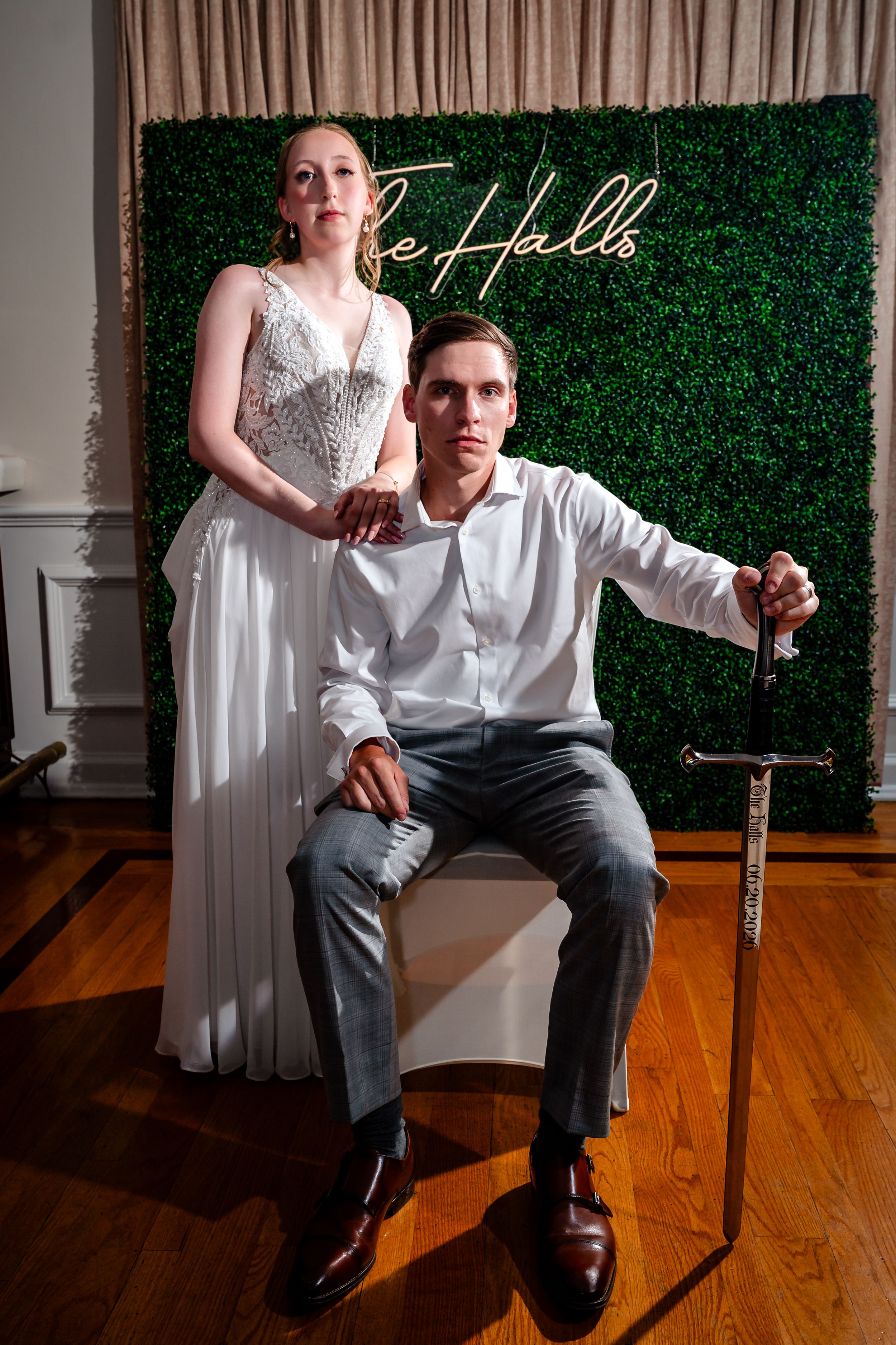 Bride and groom pose dramatically with a sword in front of a green hedge wall with neon sign.