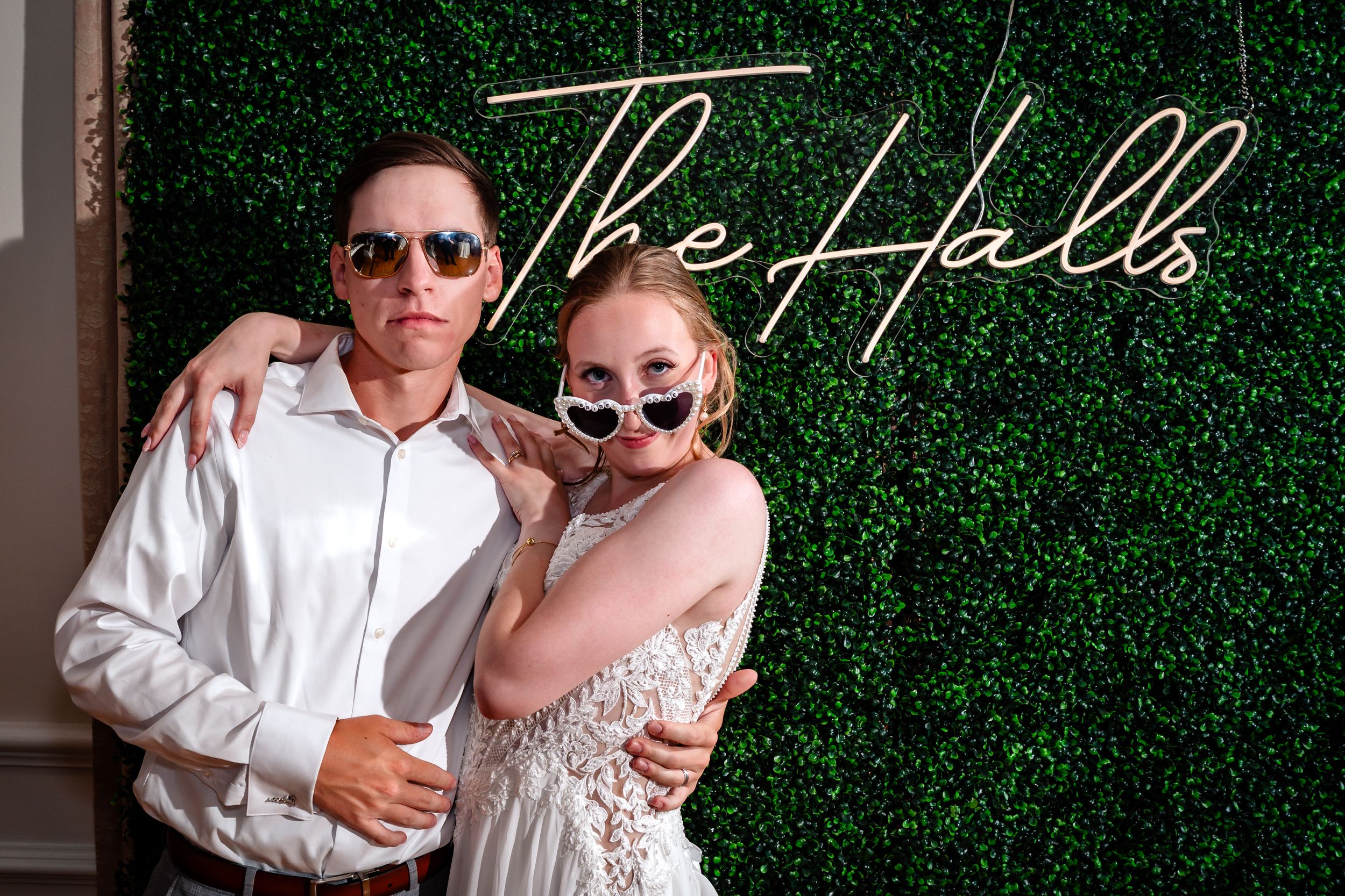 Newlyweds pose playfully with heart sunglasses at The Halls wedding photo booth backdrop.