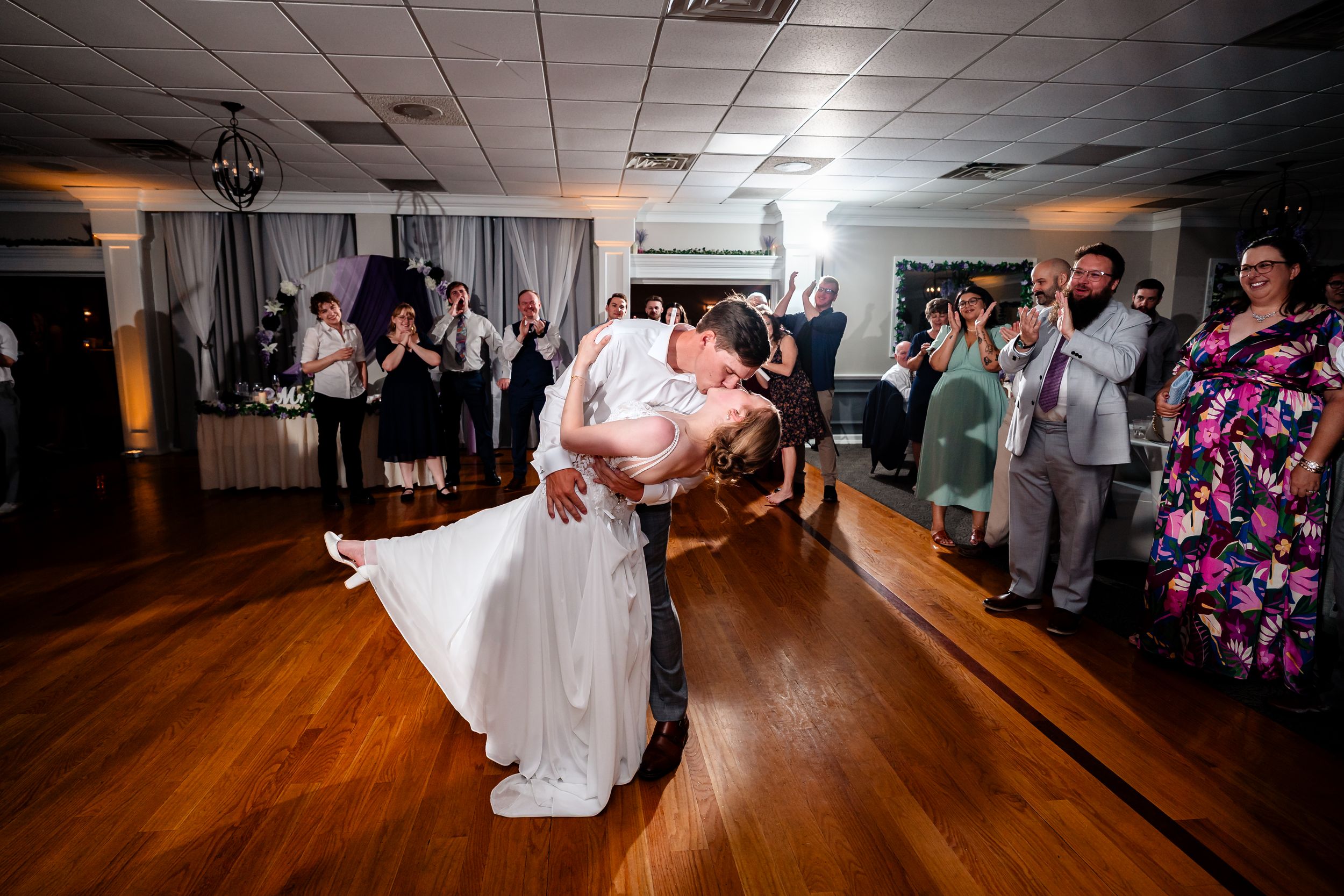 Bride and groom share romantic first dance dip at wedding reception with guests applauding in background.