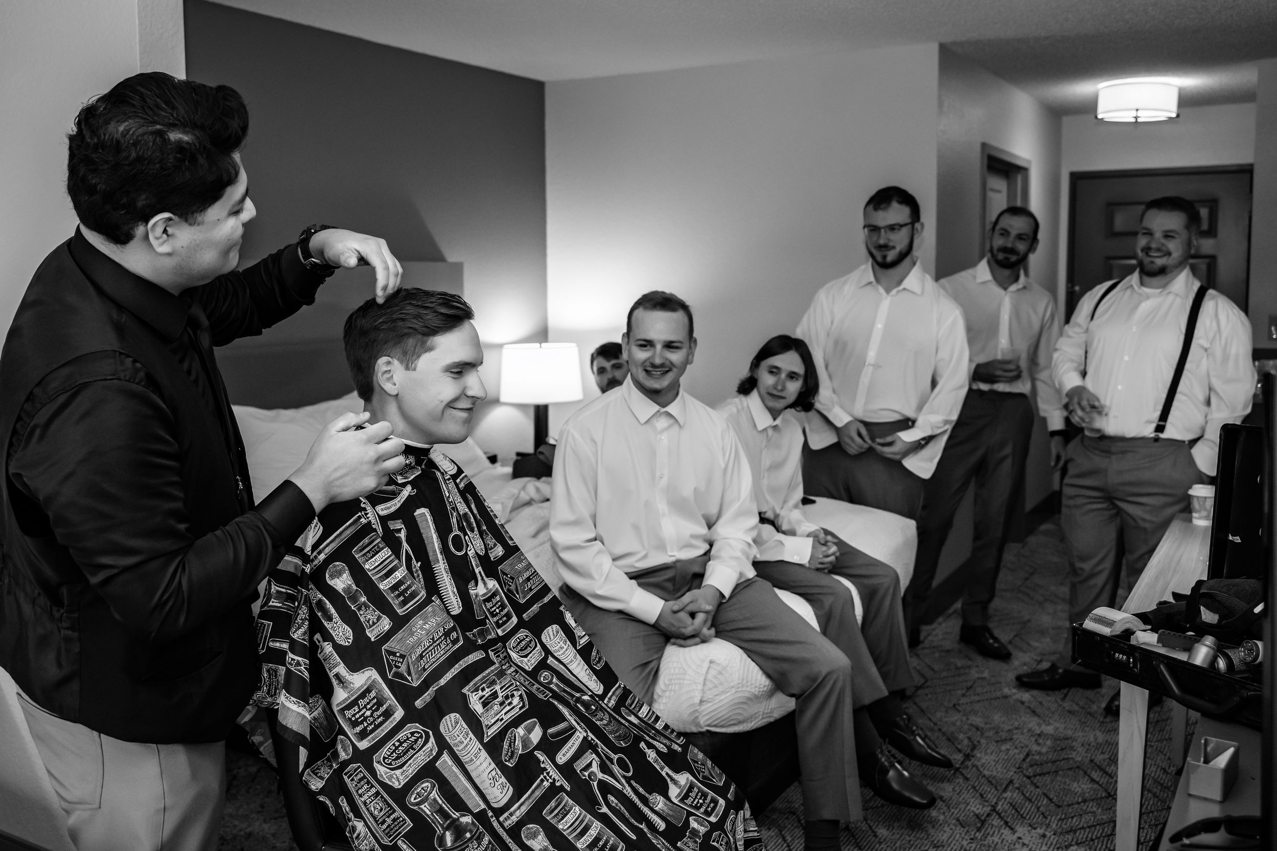 Groom gets haircut from barber while groomsmen laugh together in hotel room.
