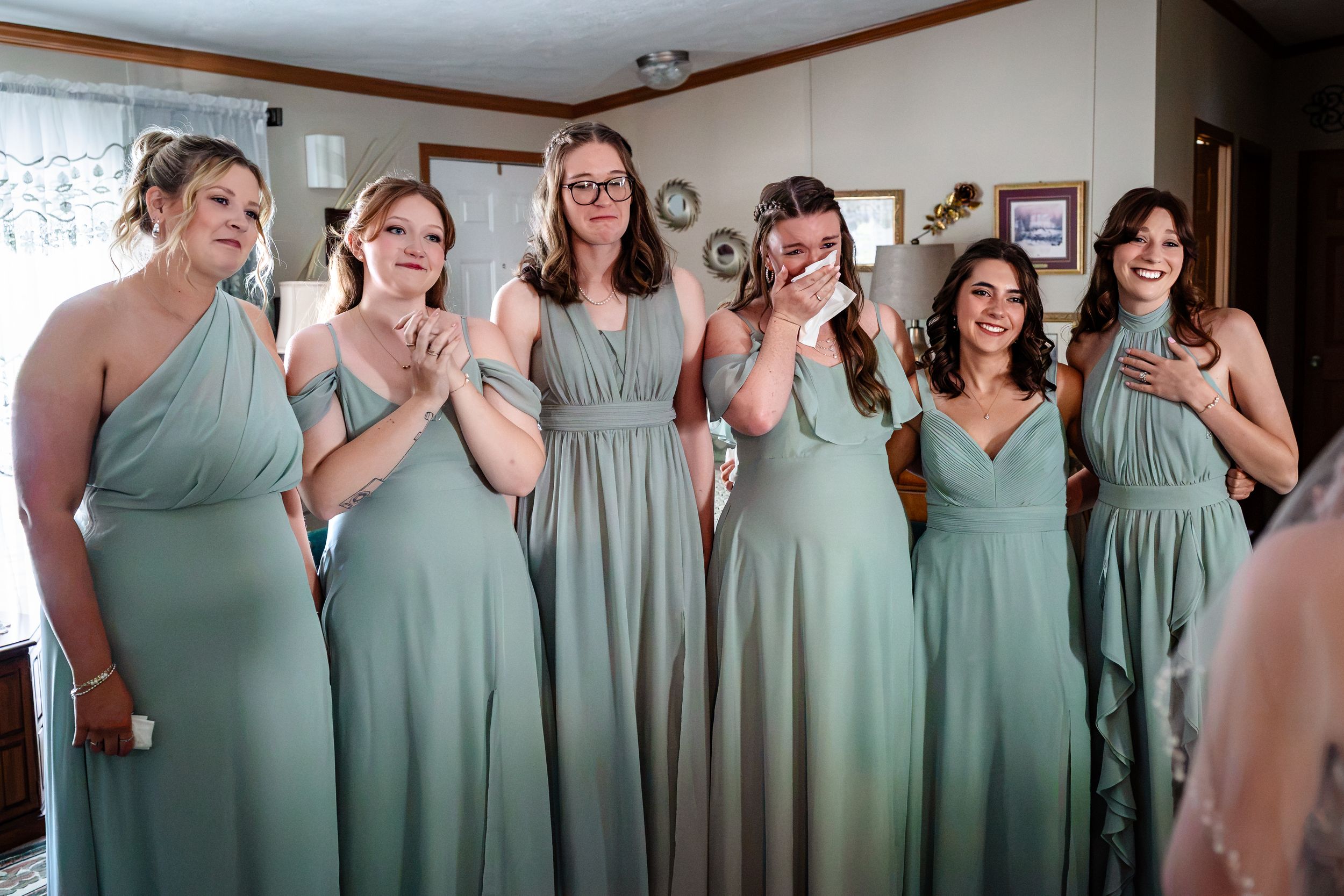 Six bridesmaids in sage green gowns reacting emotionally to bride during wedding preparations.