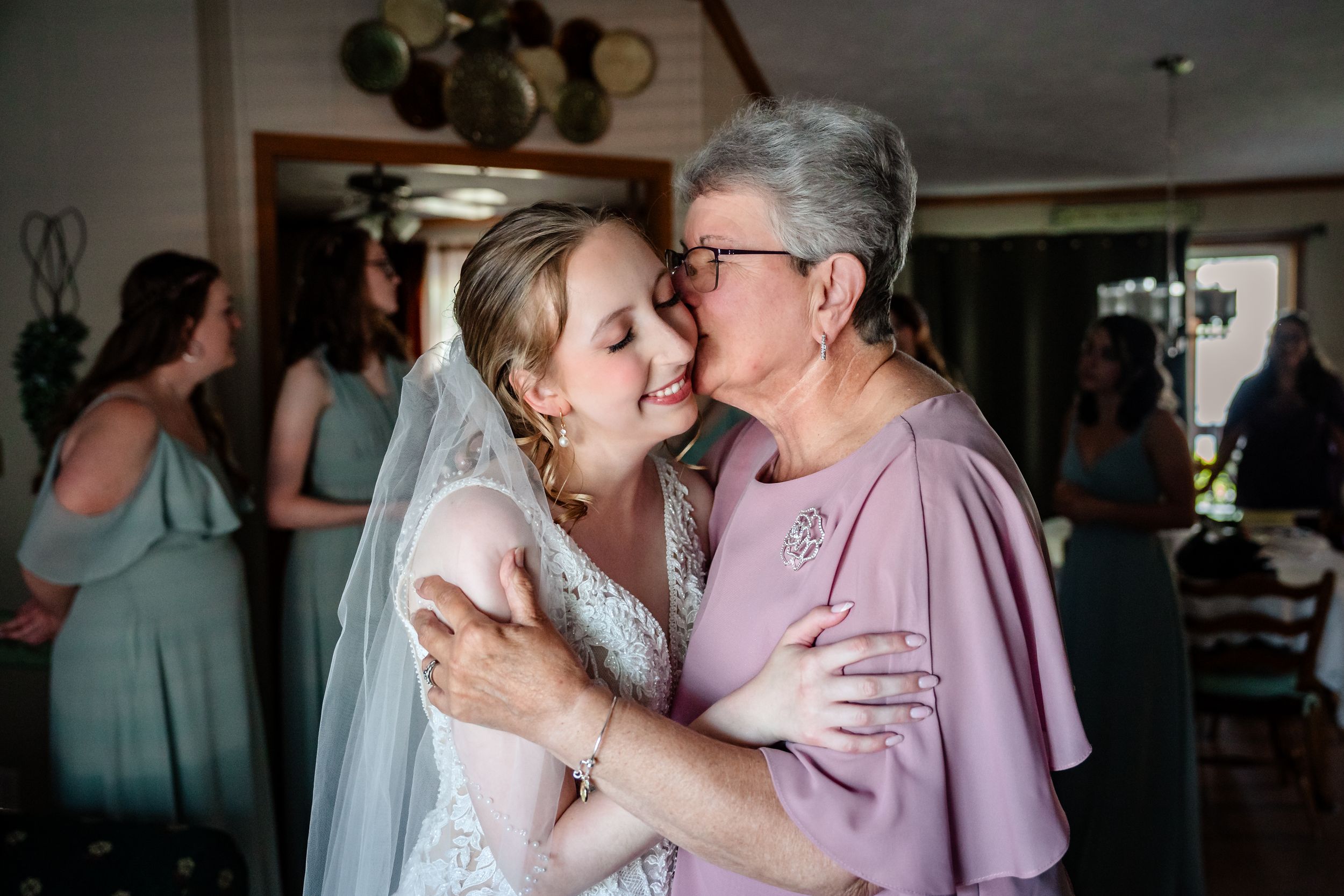 Bride in lace gown and veil shares tender embrace with older woman in lavender dress at wedding celebration.
