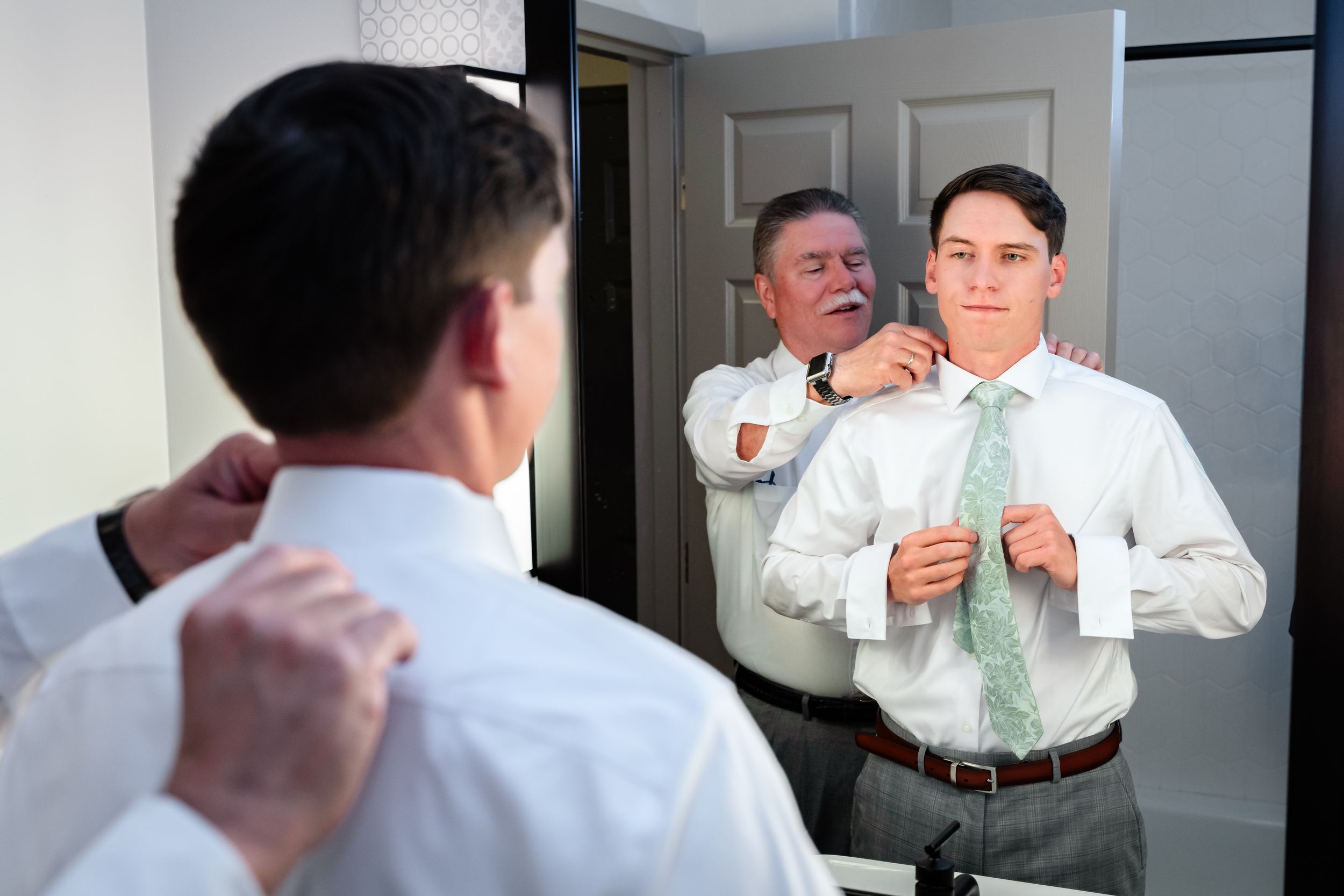 Father helping groom adjust green tie in mirror during wedding day preparations.