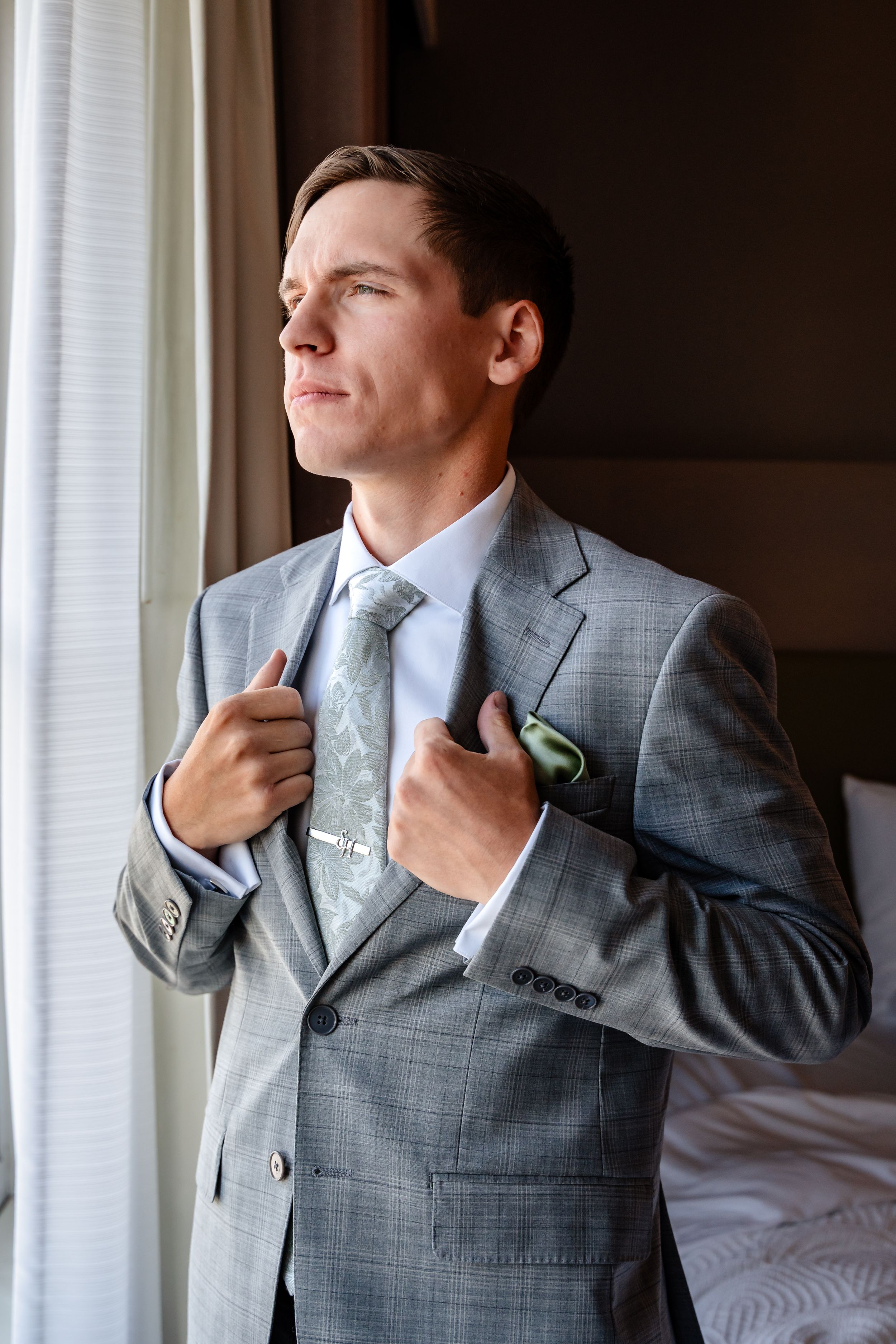 Elegant groom in grey plaid suit adjusting tie near window before wedding ceremony.