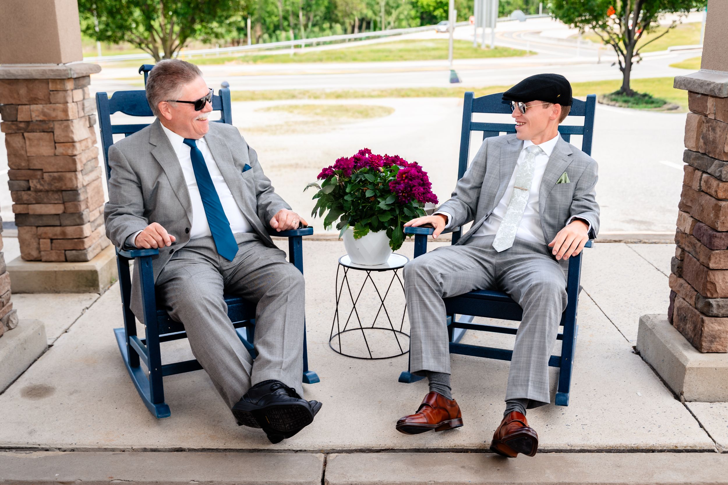 Two men in gray suits relax in blue rocking chairs on a porch before a wedding.