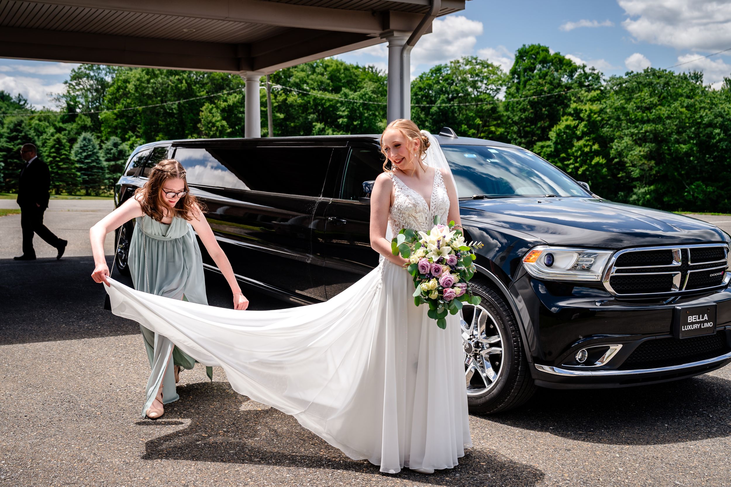 Bridesmaid helps arrange bride's white gown train beside a black SUV outdoors.