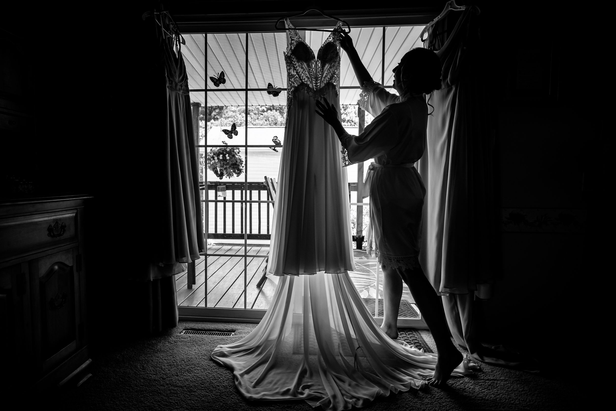Black and white silhouette of bridesmaid arranging wedding gown near a sunlit window.
