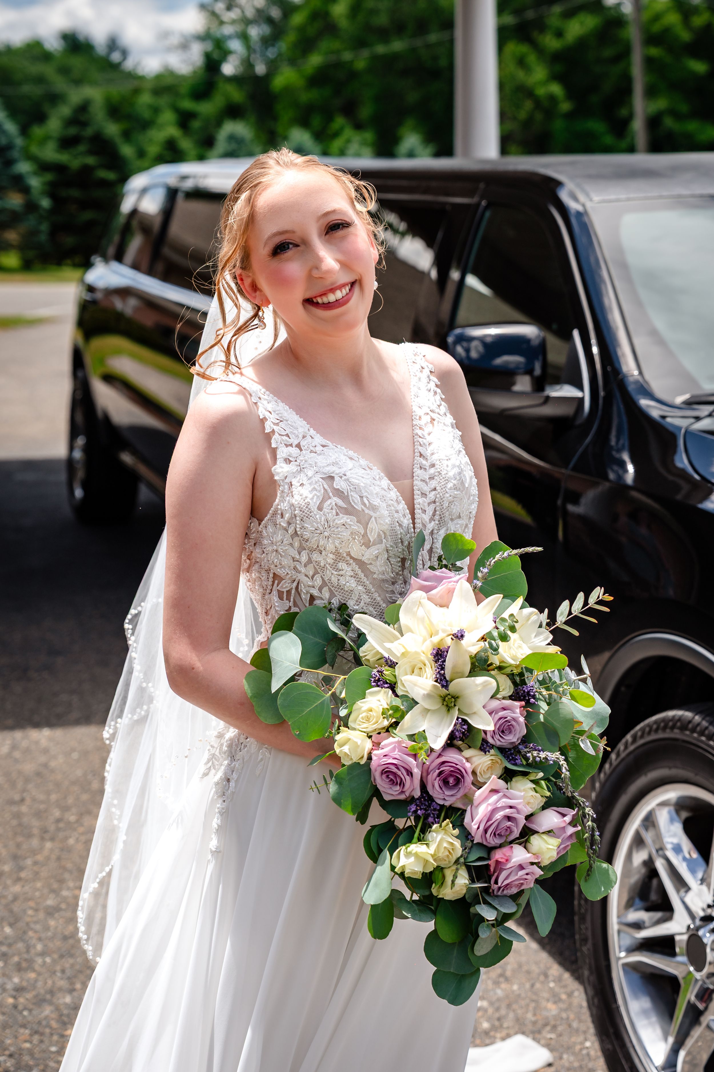 Radiant bride in lace gown holds purple and white bouquet beside a black limousine.