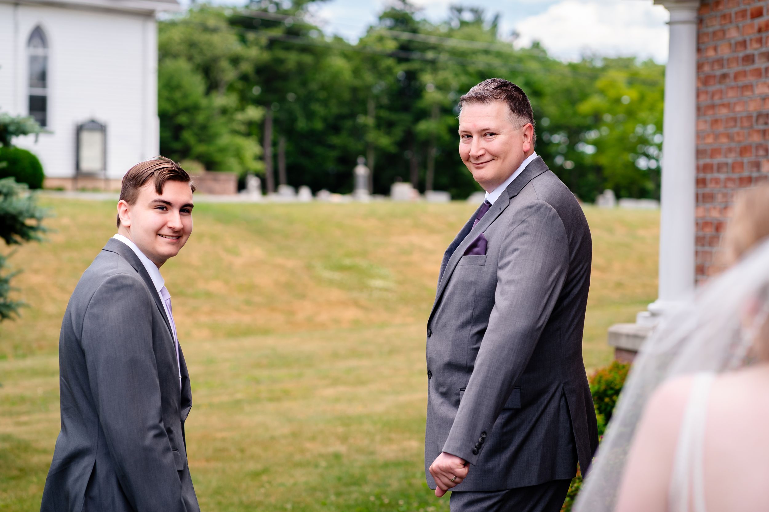 Two groomsmen in gray suits smile outdoors near a white church before wedding.