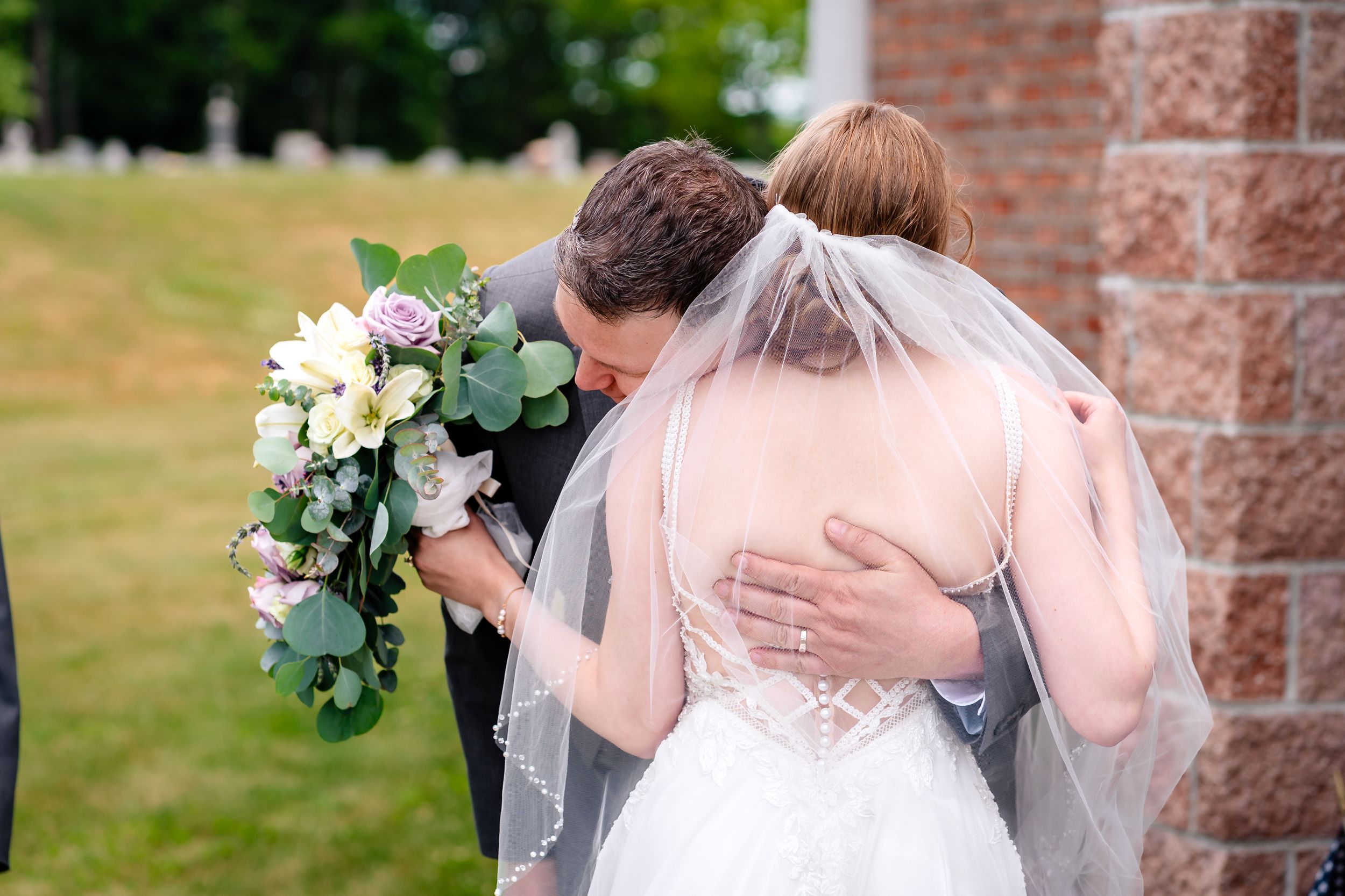 Groom embracing bride in white wedding dress and veil, holding bouquet of pink and white flowers outside brick church.