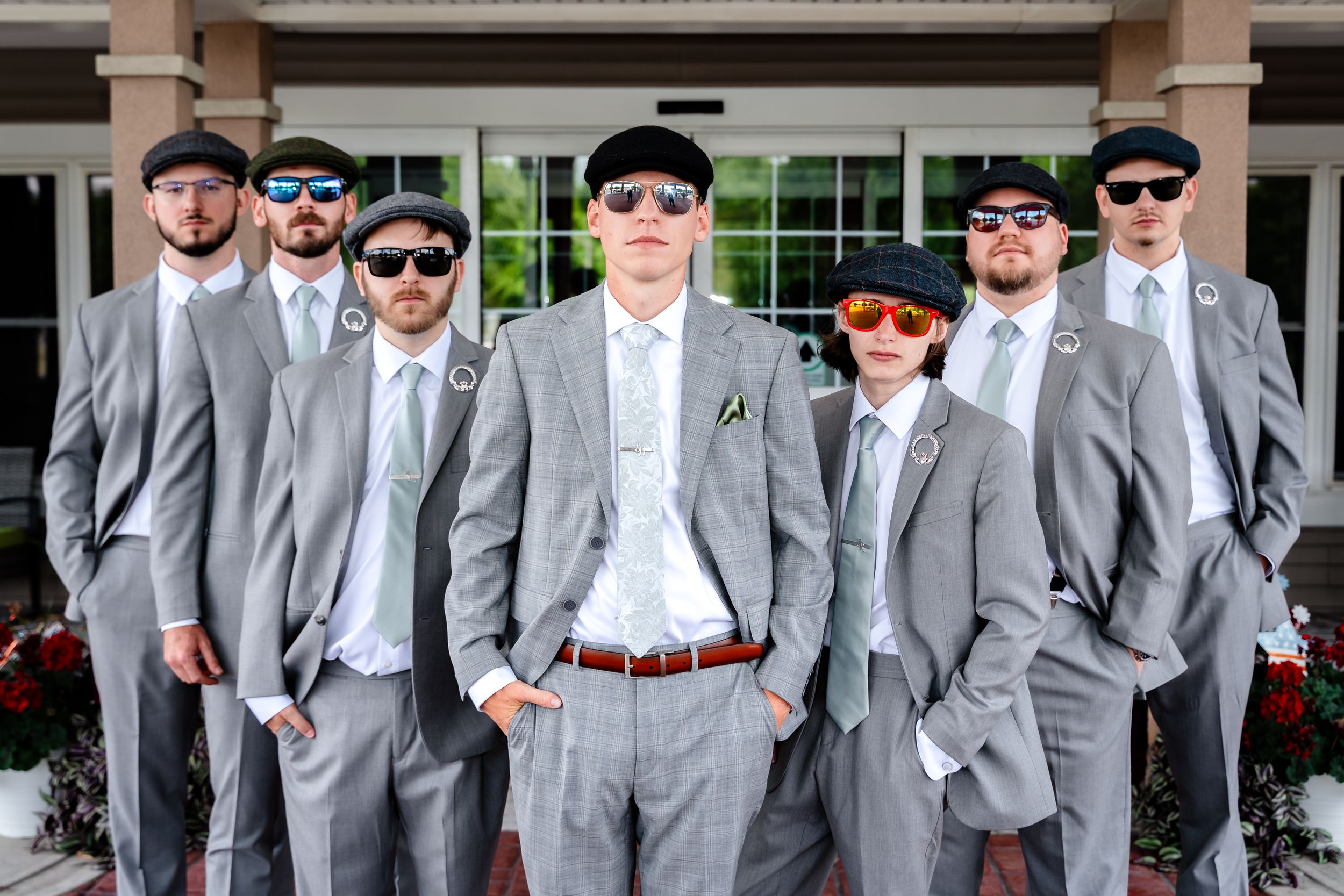 Groomsmen in matching gray suits and sunglasses pose confidently outside a venue.