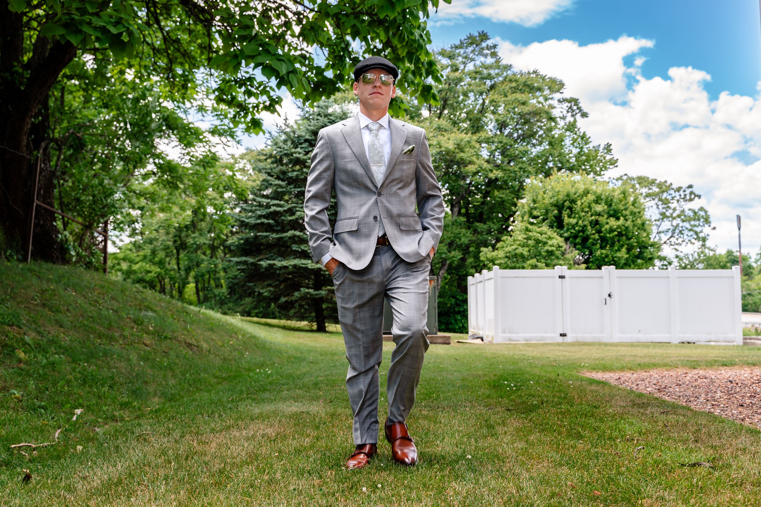 Dapper groom in gray suit and flat cap poses confidently outdoors on wedding day.