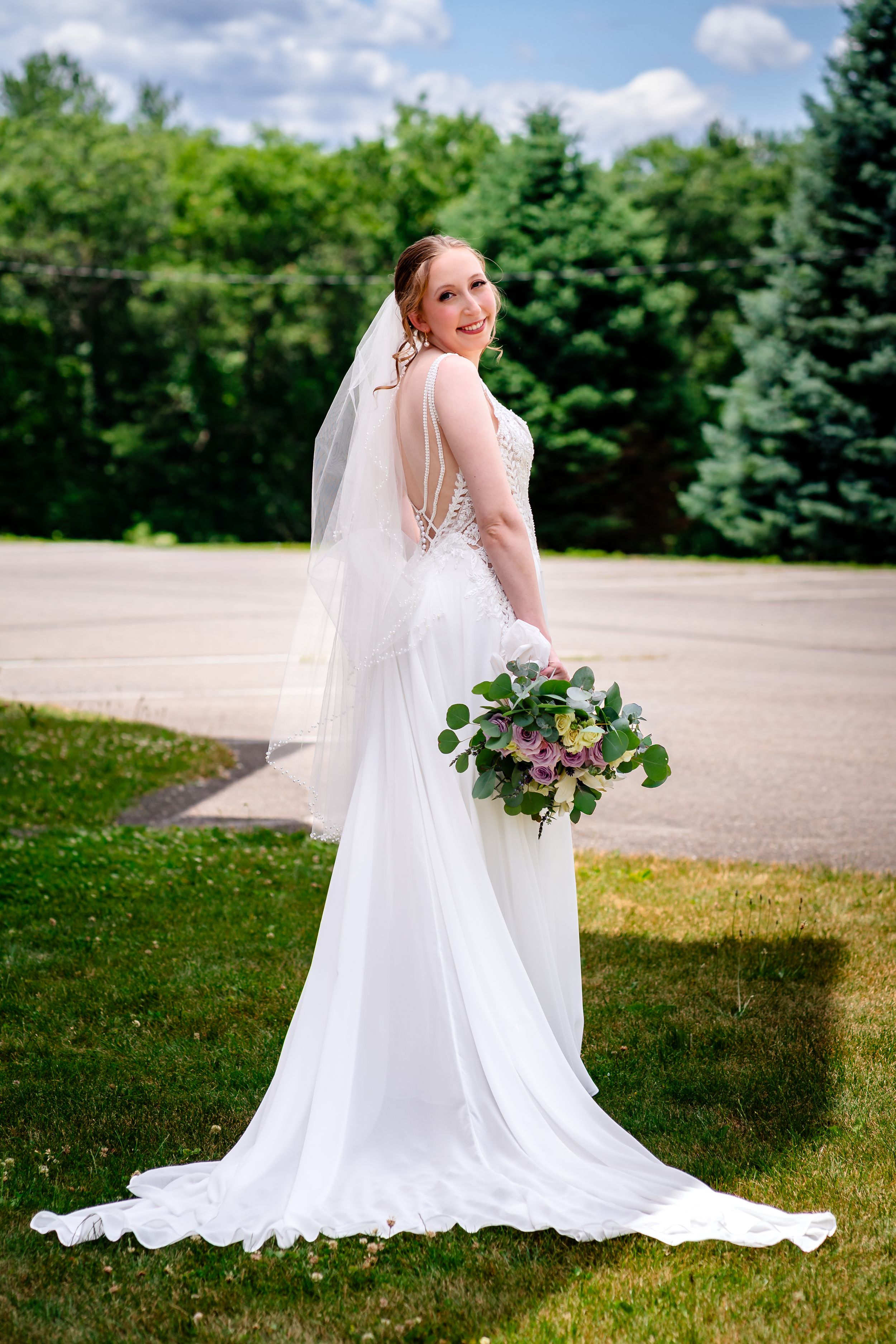 Bride in white gown and veil holds bouquet while posing outdoors on wedding day.