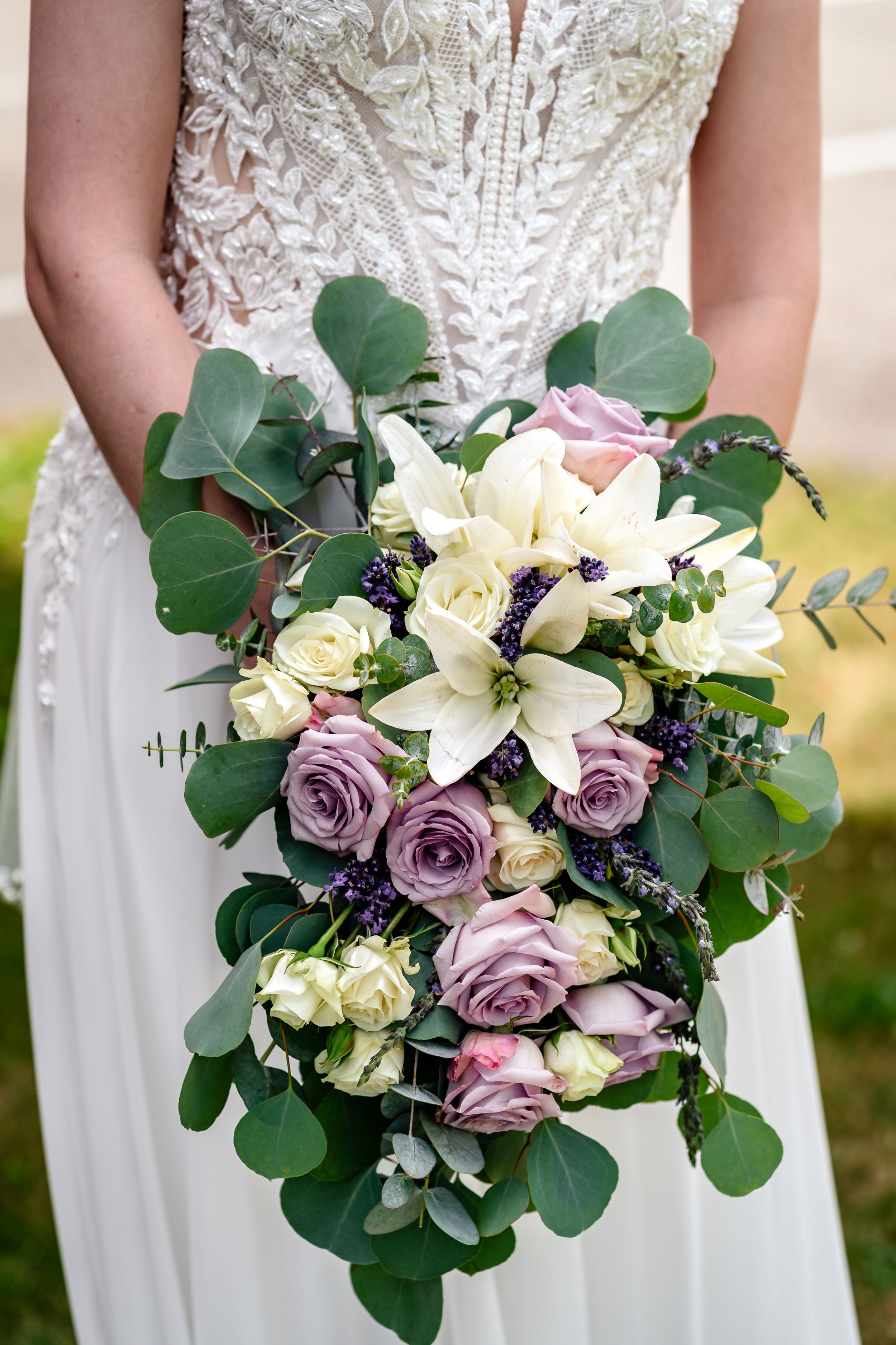 Bride holding elegant cascading wedding bouquet with white lilies, lavender roses, and eucalyptus greenery.