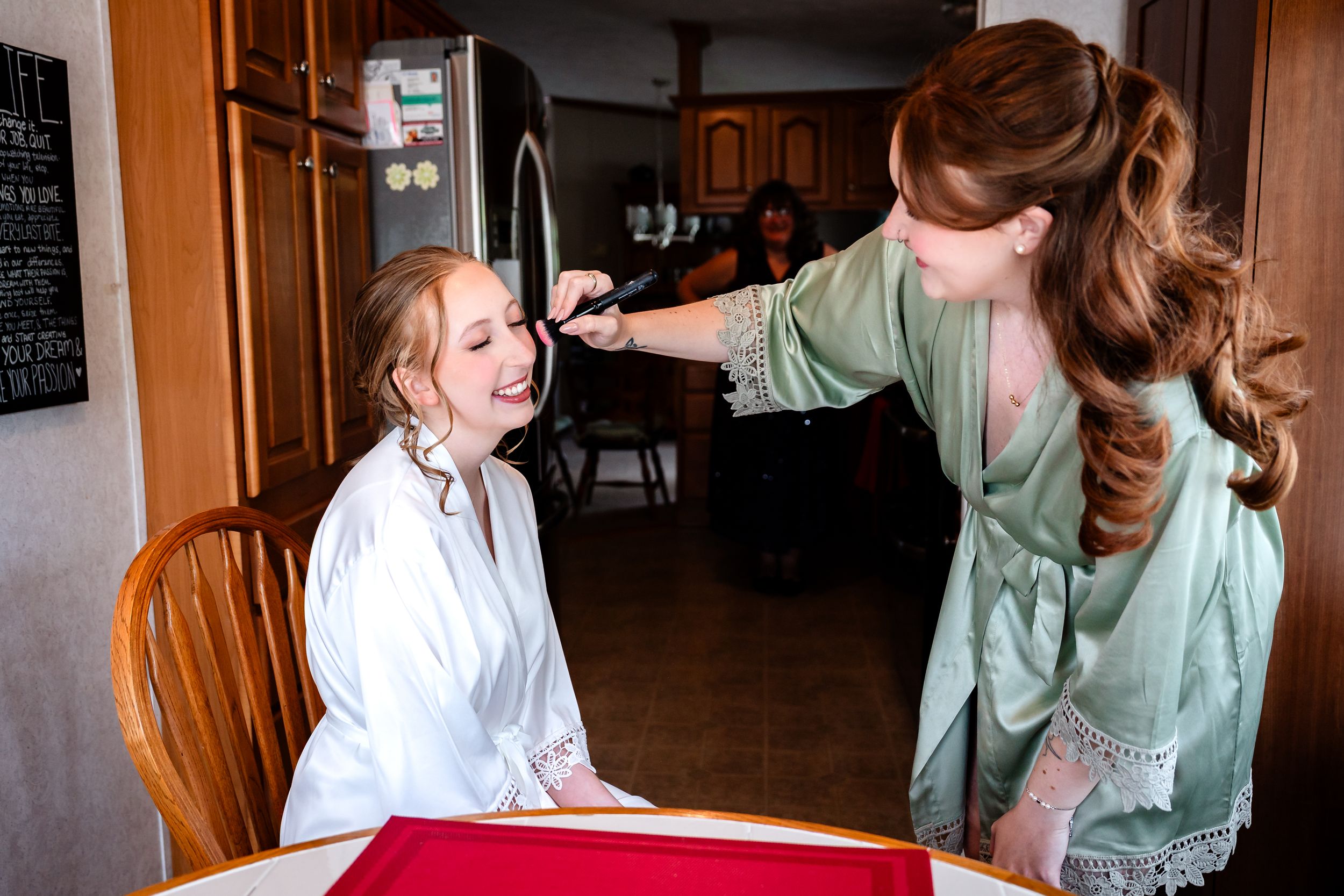 Bridesmaid applies makeup to a laughing bride in a white robe at a kitchen table.
