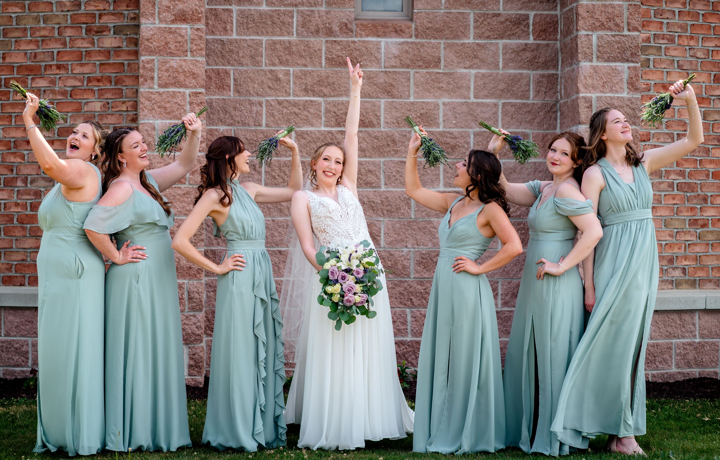 Bride poses with five bridesmaids in sage green gowns celebrating joyfully in front of brick church wall.